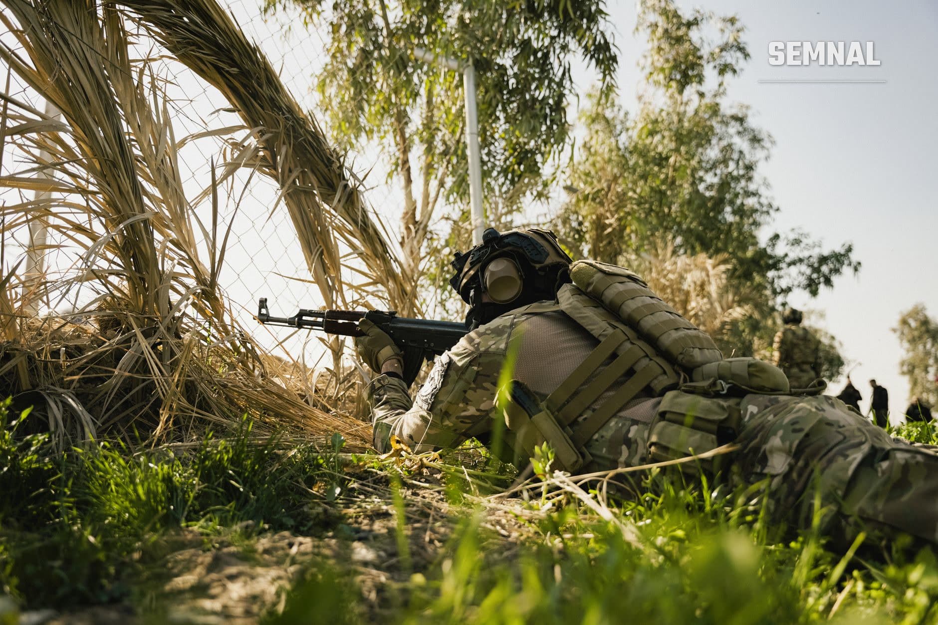 Soldat în echipament militar și masă de gaze, în poziție de tragere cu arma automată, ascuns în vegetație și teren accidentat pe front.