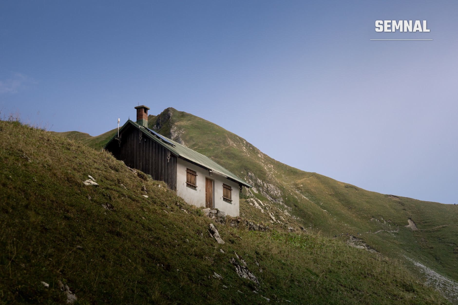 Refugiu montan tradițional cu pereți albi și acoperiș negru amplasat pe o pantă cu pajiști verzi și vegetație alpină în munți