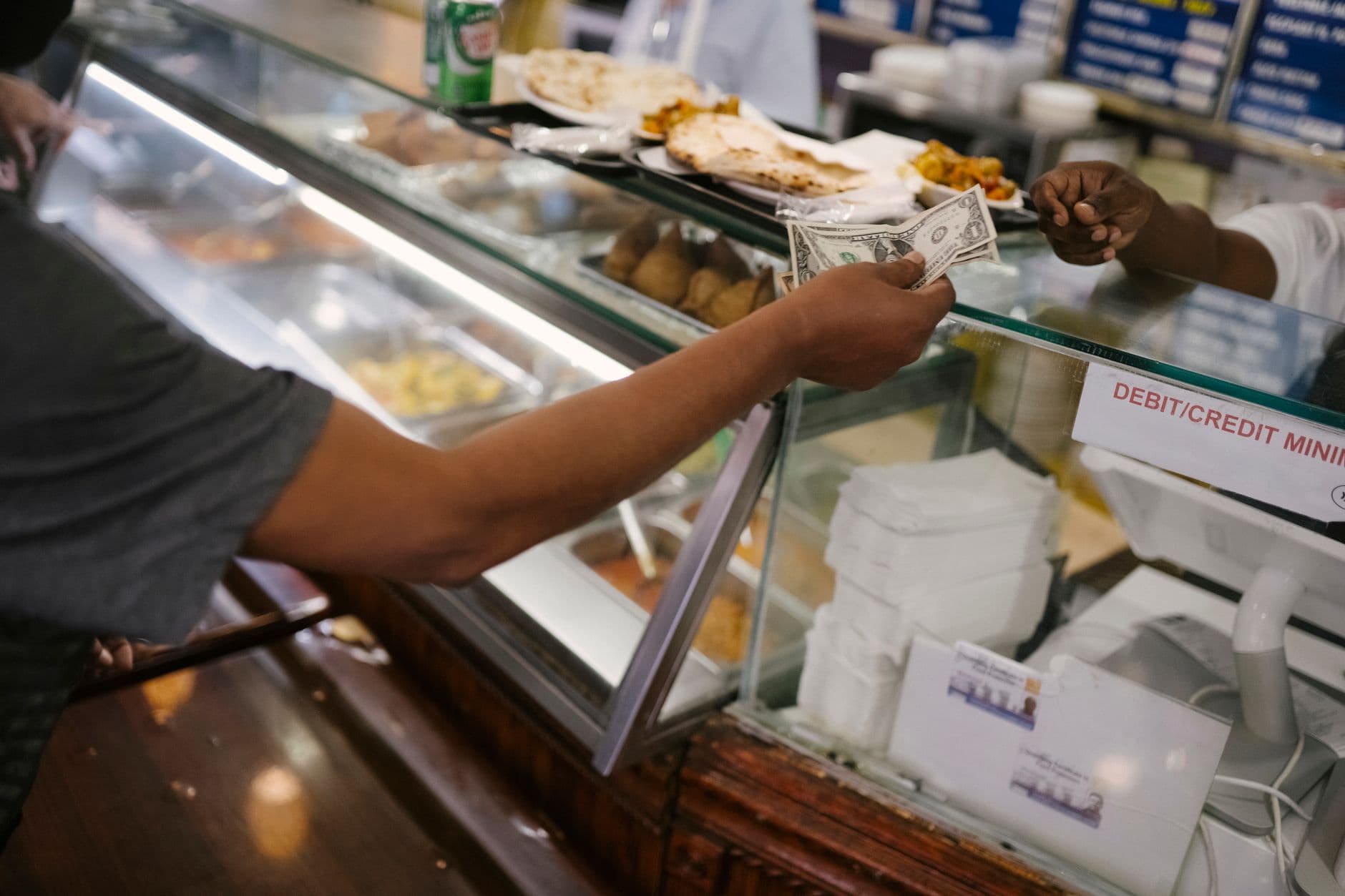 Mână care oferă o bancnotă la tejghel de fast-food, cu vitrină cu mâncare și indicator de plată cu card în fundal