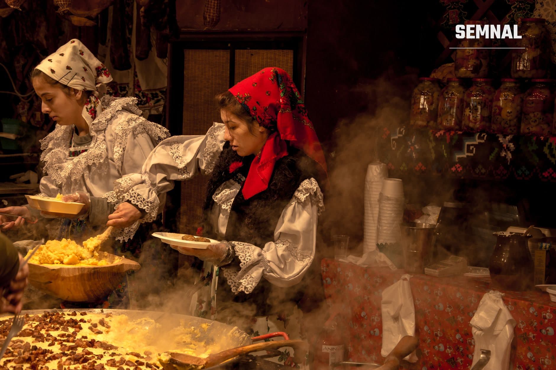 Două femei în costume și cămăși tradiționale românești cu dantele, în cămări rustic ornamentate, pregătesc mâncare cu concentrare în vase de lut cu aburi denși.