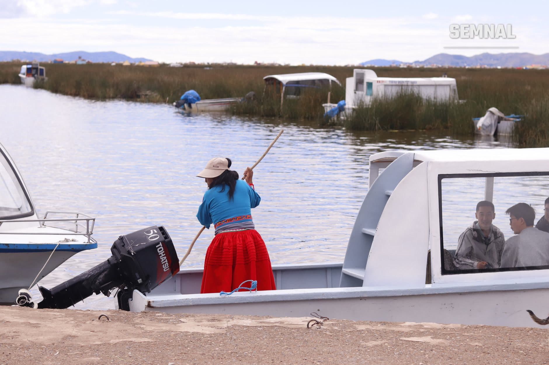 Femeie din comunitatea Uros pe lacul Titicaca, îmbrăcată în haine tradiționale, navigând barca din trestie cu pagaia; în fundal, casele plutitoare și alte bărci ale satului.