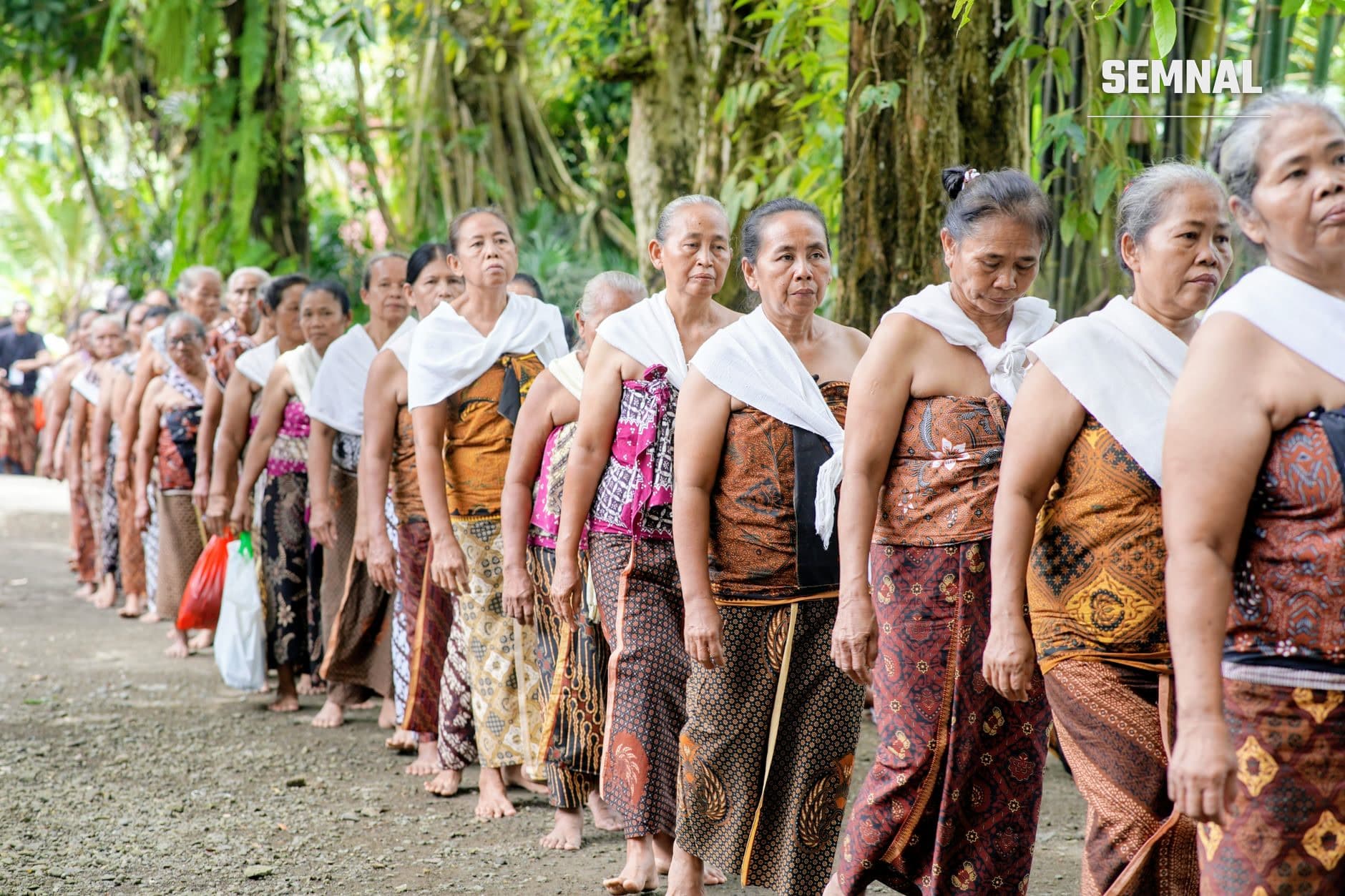 Rând de oameni din Toraja purtând îmbrăcăminte tradițională cu modele batik și șaluri albe, sub vegetație tropicală deasă, participând la ritual comunitar solemn (Foto: Ruyat Supriazi / Pexels)