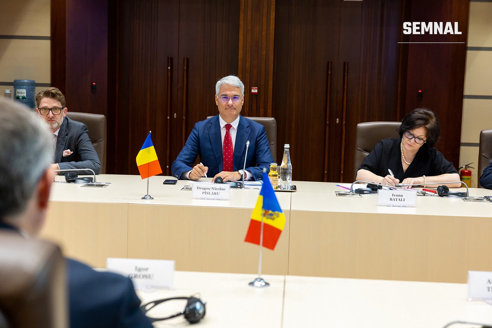 Dragoș Pîslaru, bărbat cu părul alb și ochelari, purtând costum bleumarin și cravată roșie, șezând la o masă de conferință lângă steaguri ale României, în mod oficial și formal. (Foto: Parlamentul Republicii Moldova | Pagina oficială)