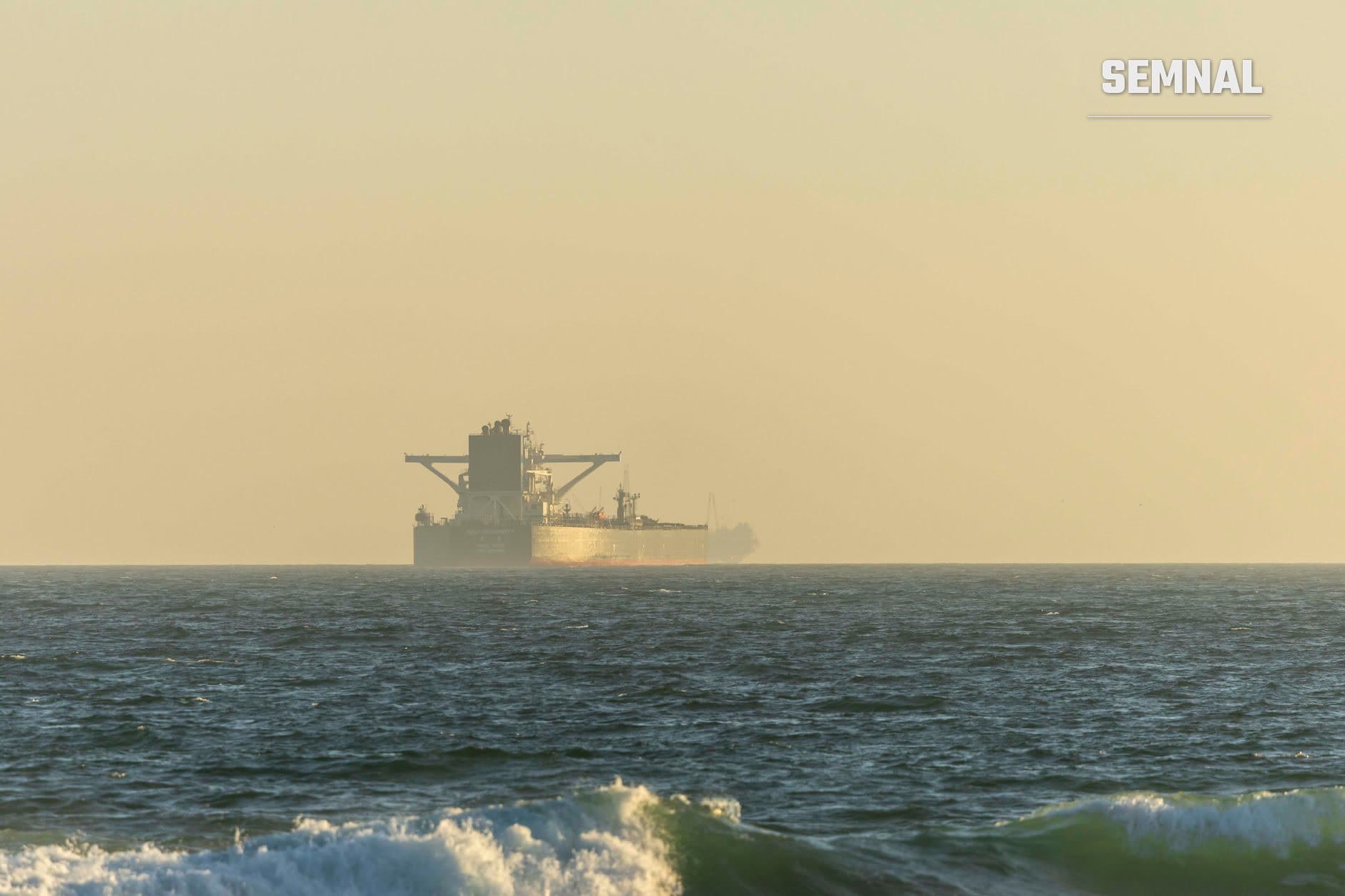 Navă petrolier de mari dimensiuni ancorată în larg, fotografiată de pe plajă cu valuri în prim-plan și cer deschis pe apus. (Foto: Stephen Leonardi / Pexels)