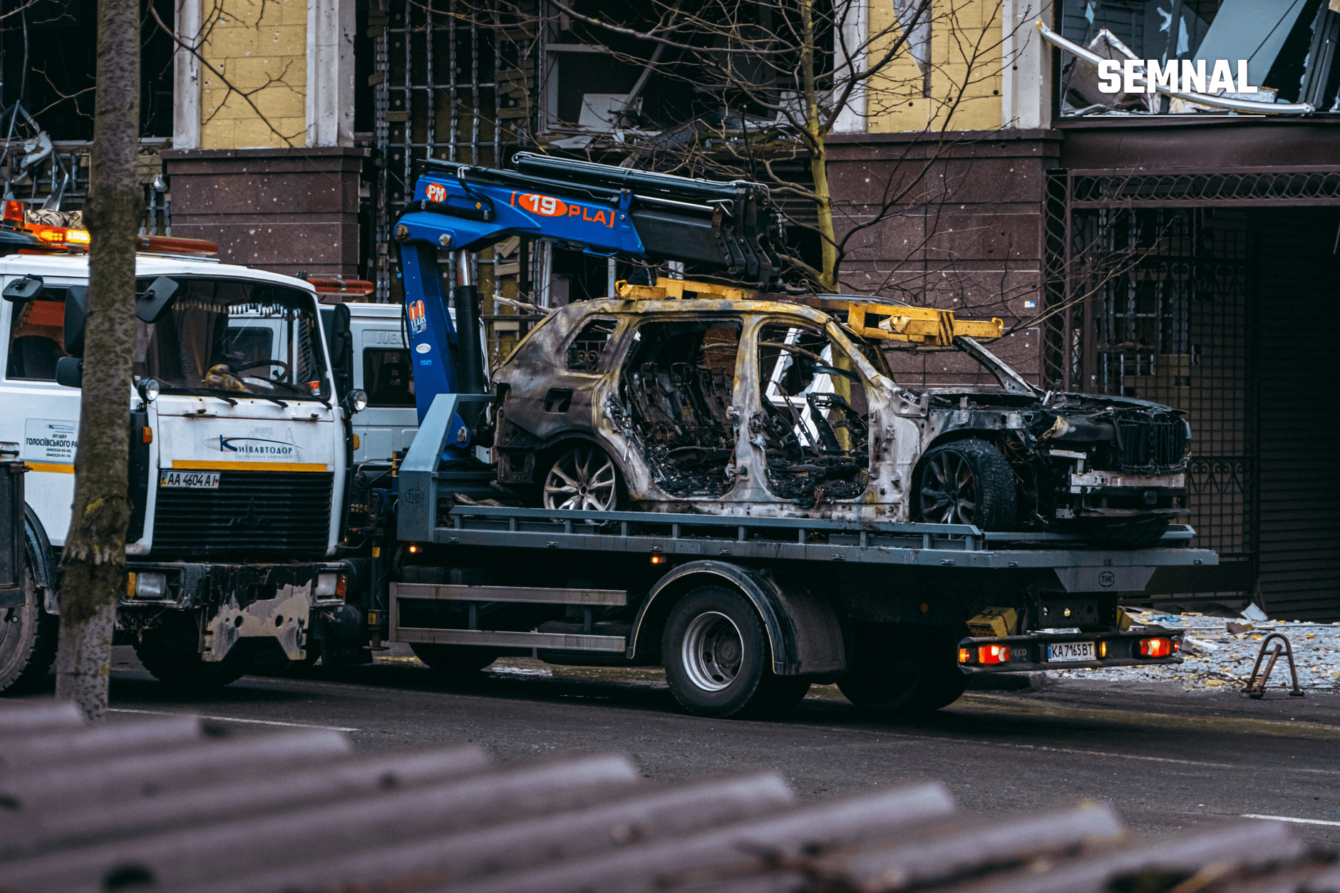 Vehicul complet distrus și carbonizat încărcat pe o platformă de remorcare în centrul unui oraș urban, cu clădiri comerciale și echipamente de intervenție în prim plan. (Foto: Алесь Усцінаў / Pexels)