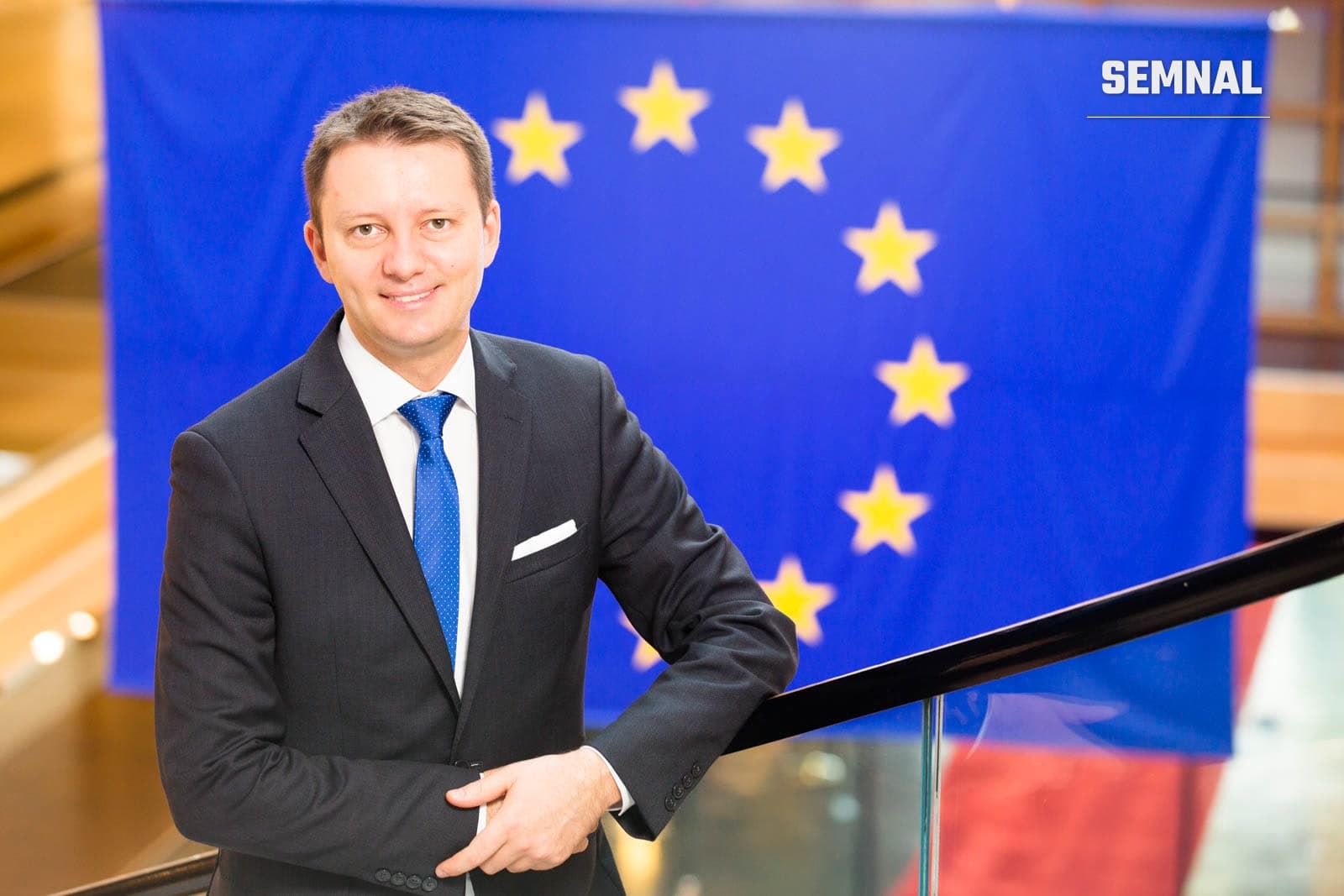 Siegfried Mureșan, bărbat în costum negru și cravată albastră, pozat în fața steagului Uniunii Europene cu stele galbene pe fundal albastru. (Foto: AureliaA)