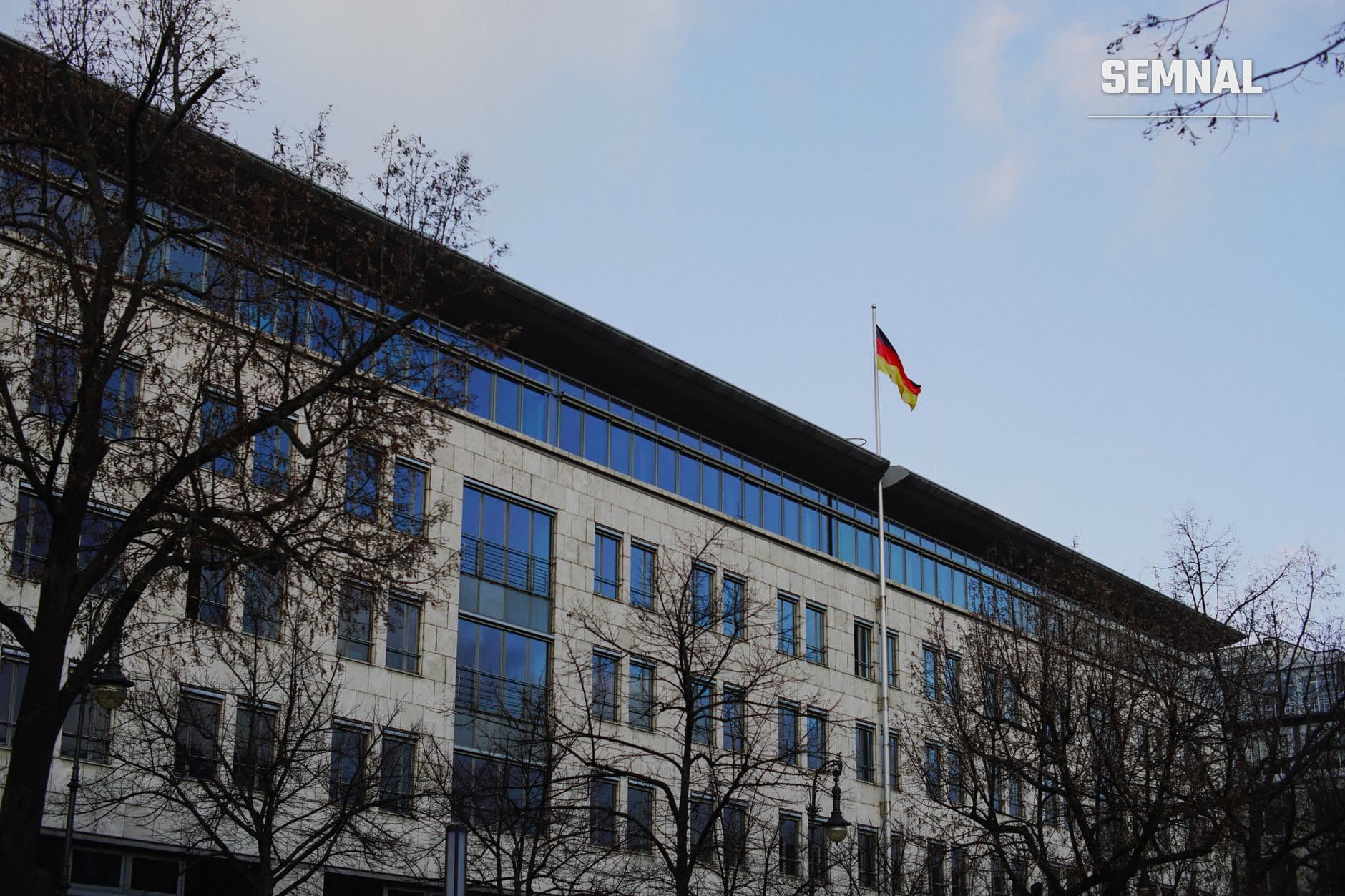 Clădire oficială germană cu fațadă din piatră albă și ferestre albastre, cu drapelul Germaniei pe catarg, reprezentând instituția diplomatică care se închide. (Foto: Cristiano Junior / Pexels)