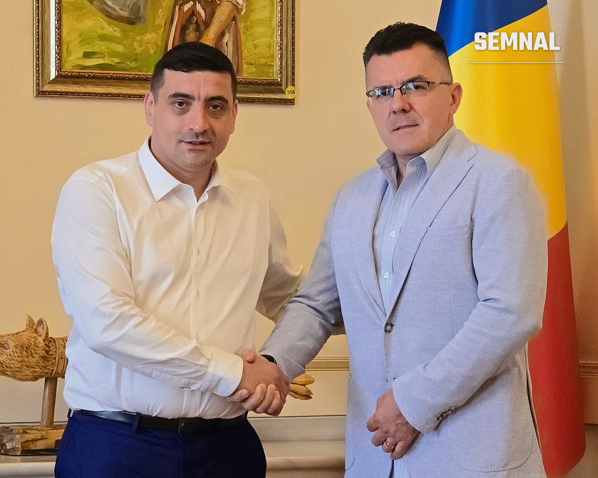 George Simion, în cămașă albă și pantaloni bleumarin, salută cu strângere de mână un bărbat în sacou gri și ochelari, într-o încăpere oficială cu drapelul României în fundal. (Foto: AUR Alianța pentru Unirea Românilor)