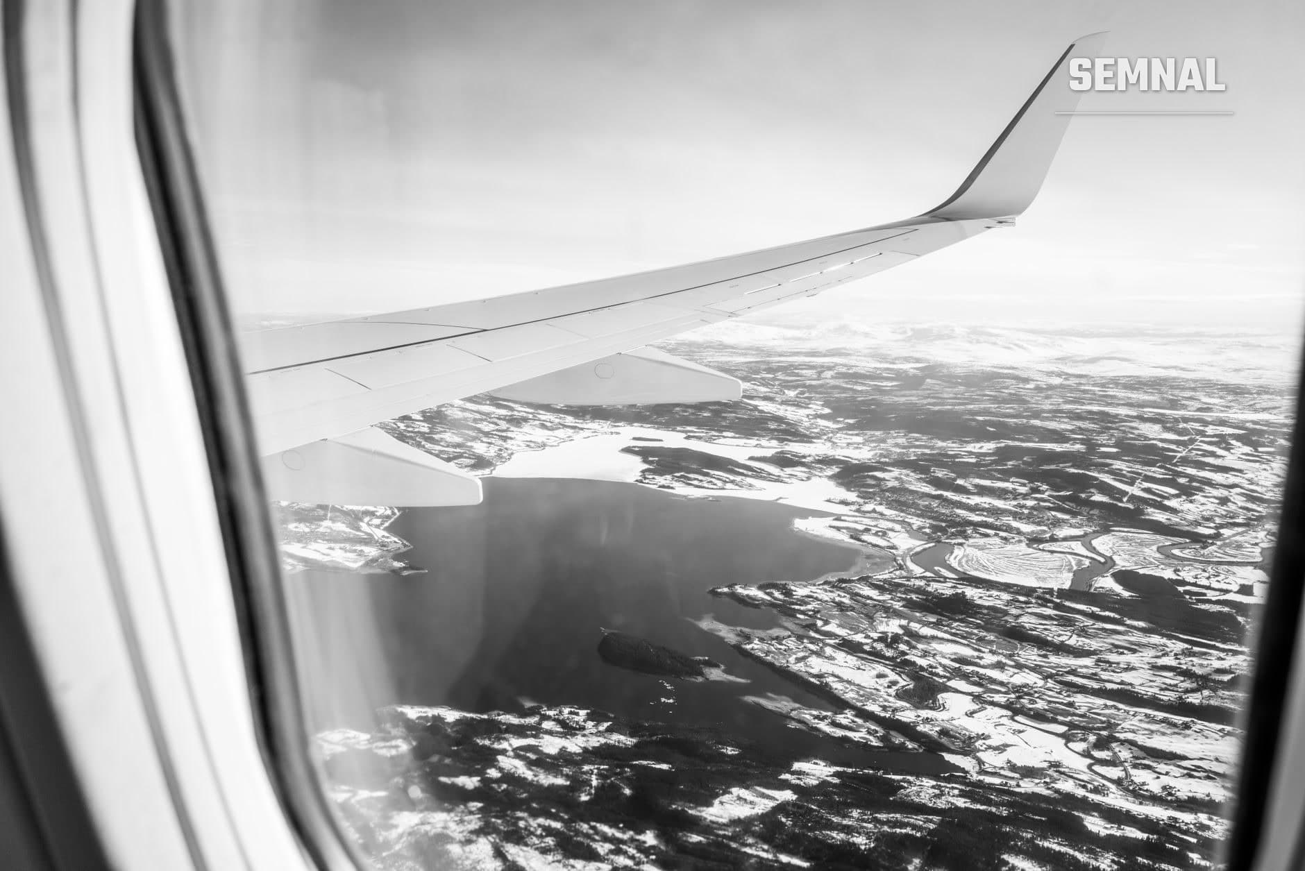 Vedere din fereastră de avion în alb-negru: ala avionului și peisaj montan acoperit de zăpadă cu lacuri și păduri în sezonul de iarnă. (Foto: Boris K. / Pexels)