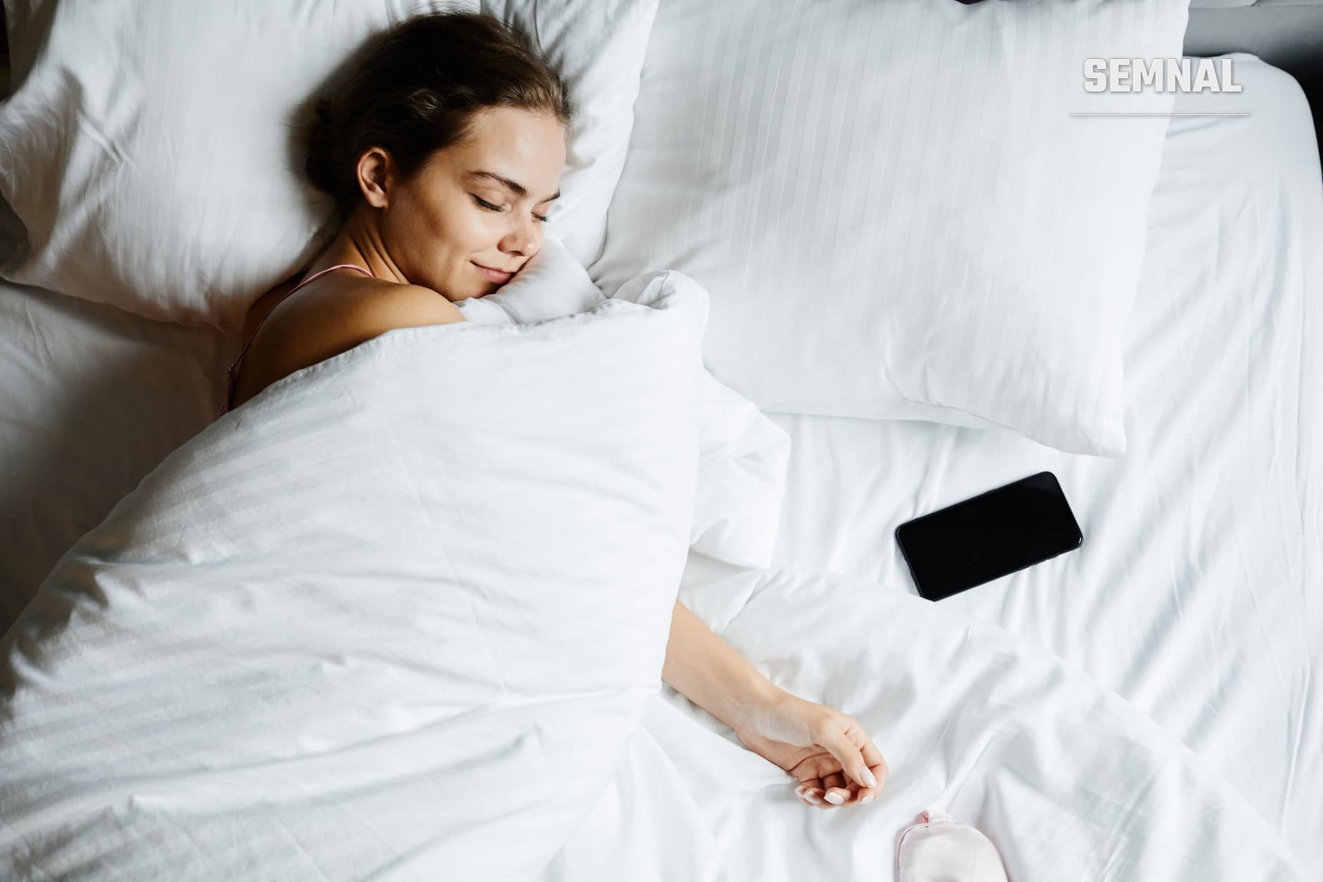 Femeie care doarme pașnic pe pernă albă în pat, cu un telefon mobil negru lângă ea, ilustrând importanța igienei somnului și influența dispozitivelor electronice asupra odihnei (Foto: Marcus Aurelius / Pexels)
