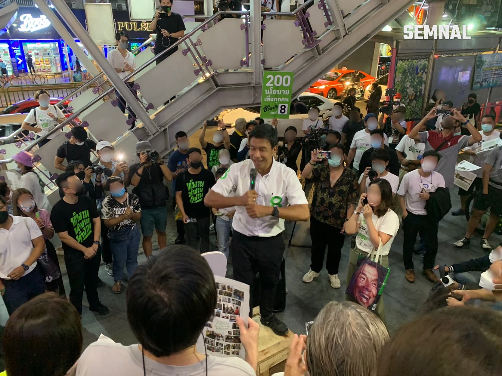 Chadchart Sittipunt, guvernatorul orașului Bangkok, în cămașă albă, în prim-plan la un eveniment public într-un centru comercial, înconjurat de mulțime și jurnaliști. (Foto: Chainwit.)