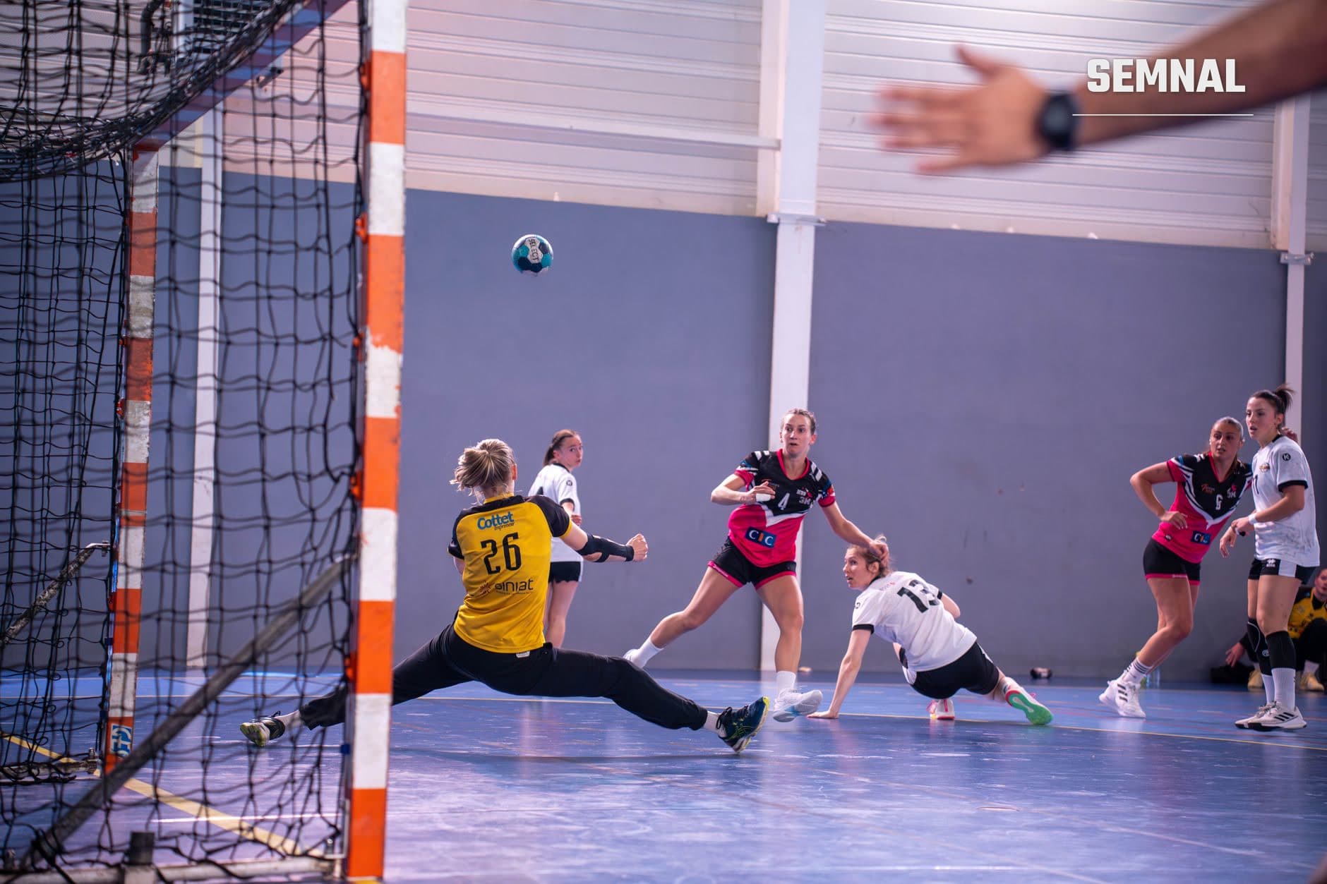 Jucătoare de handbal în tricou galben apărând la poartă cu stâlp portocaliu-alb, în meci oficial feminin pe teren albastru, cu adversare în echipamente roșu-negru și alb. (Foto: Laura Rieusset / Pexels)