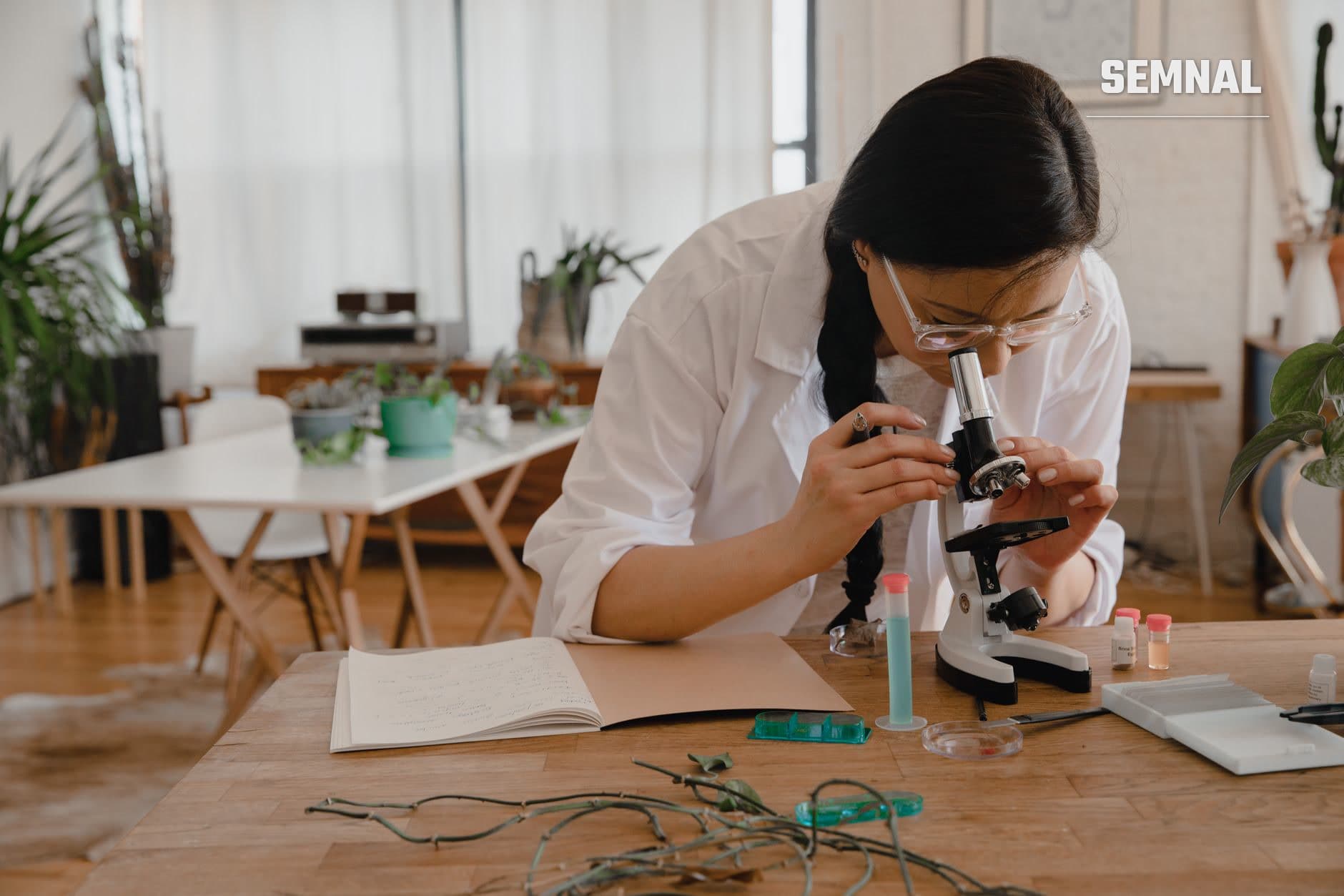Cercetătoare în halat alb lucrând la microscopul optic cu eșantioane în laborator luminos, având pe masă caiete și echipament de cercetare științifică. (Foto: RDNE Stock project / Pexels)