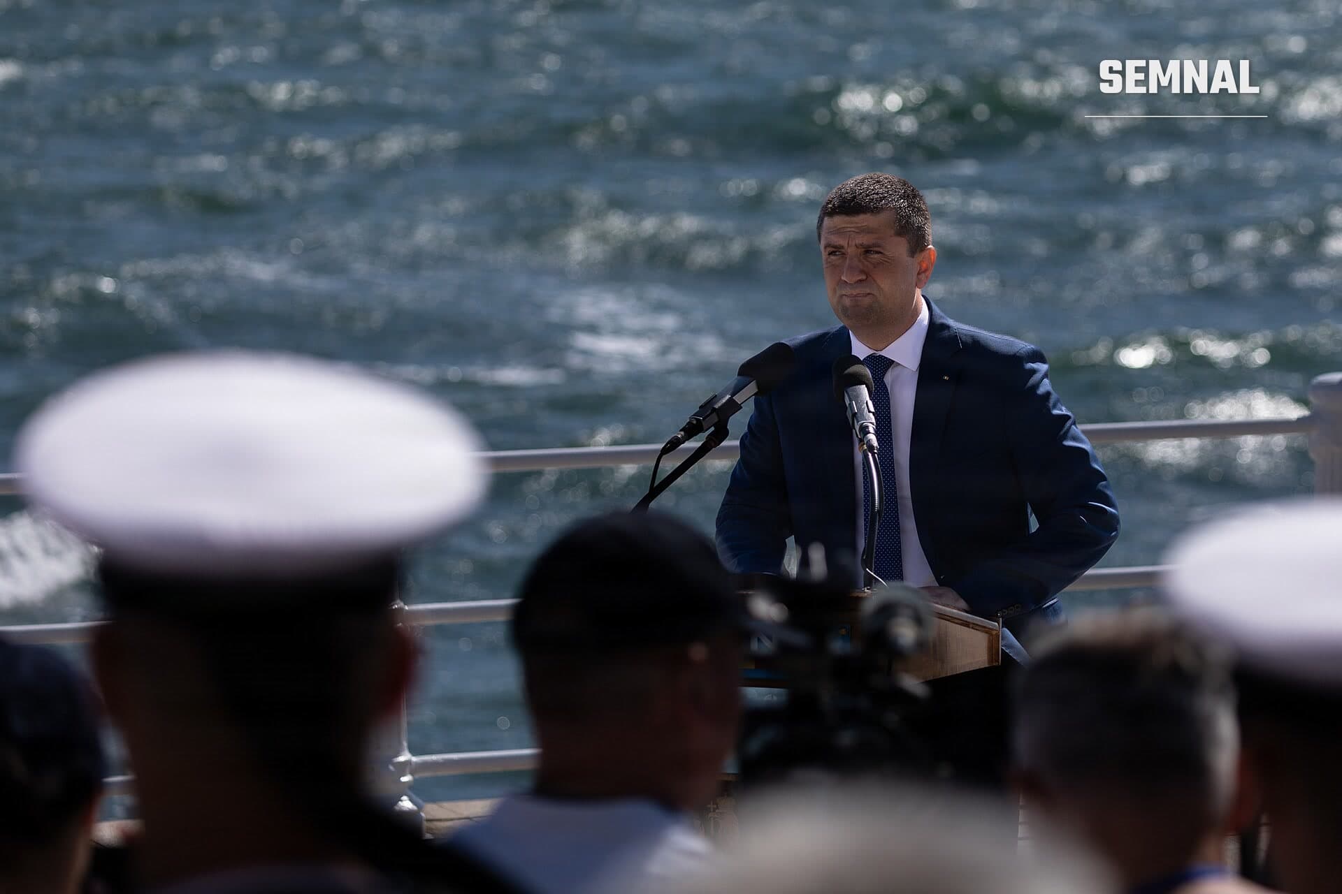 Radu Miruță vorbind la podium în fața unui grup de oameni, purtând costum bleumarin, cu apă în fundal. (Foto: U.S. Army Reserve photo by Sgt. Alex Romanack)
