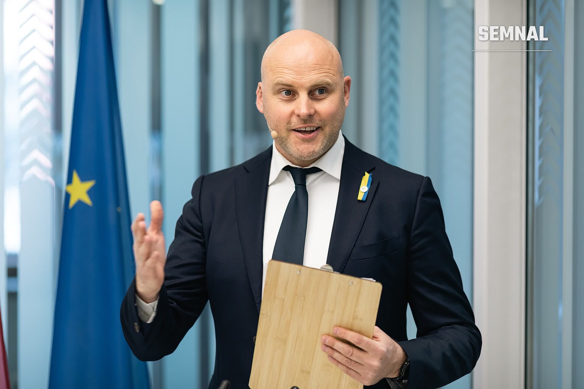 Andris Kulbergs, prim-ministru al Letoniei, ținând un dosar din lemn în fața unei drapele europene, într-o fotografie de presă din clădire cu pereți din sticlă. (Foto: Saeima)