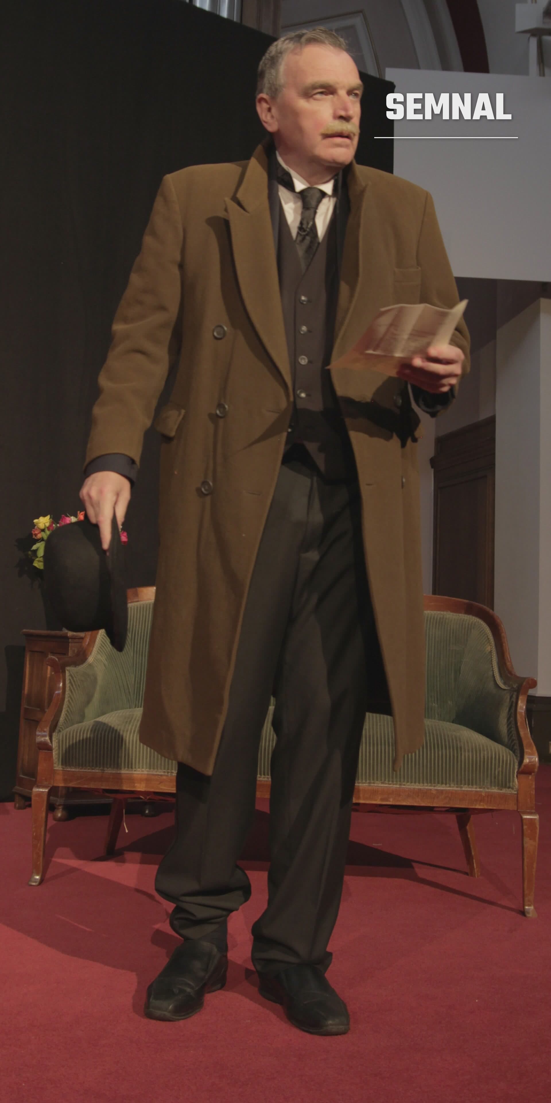 David Robinson în costum maro și vest negru, ținând o floare și un document, la un eveniment indoor pe podium. (Foto: Viggosway)
