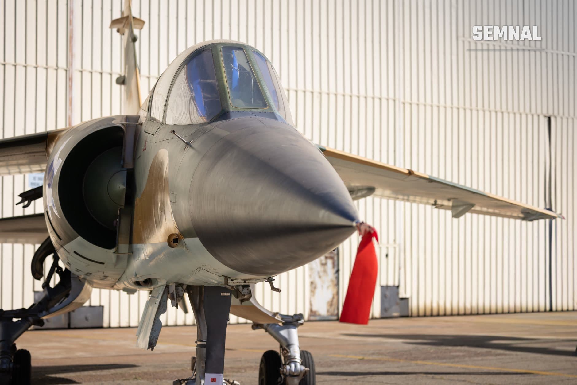 Bombardier militar modern cu fuselaj gri și cabină albastră parcat în hangarul unei baze aeriene, cu ușă roșie de hangar în fundal (Foto: Magda Ehlers / Pexels)