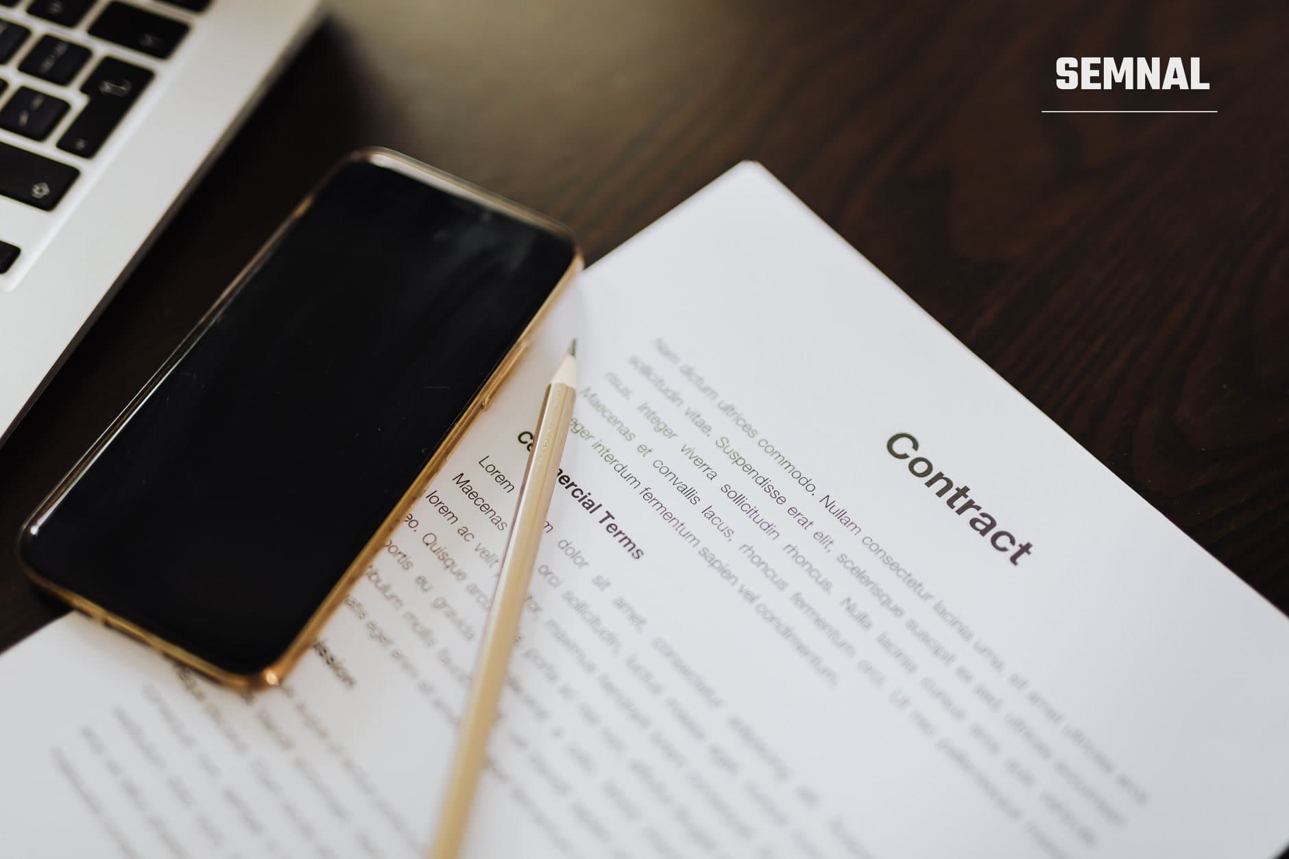 Document contract deschis pe masă de birou cu titlul 'Contract' și clauze imprimateVisibil, alături de un telefon mobil auriu și un creion, într-o scenă de verificare înainte de semnare. (Foto: https://kaboompics.com/ / Pexels)