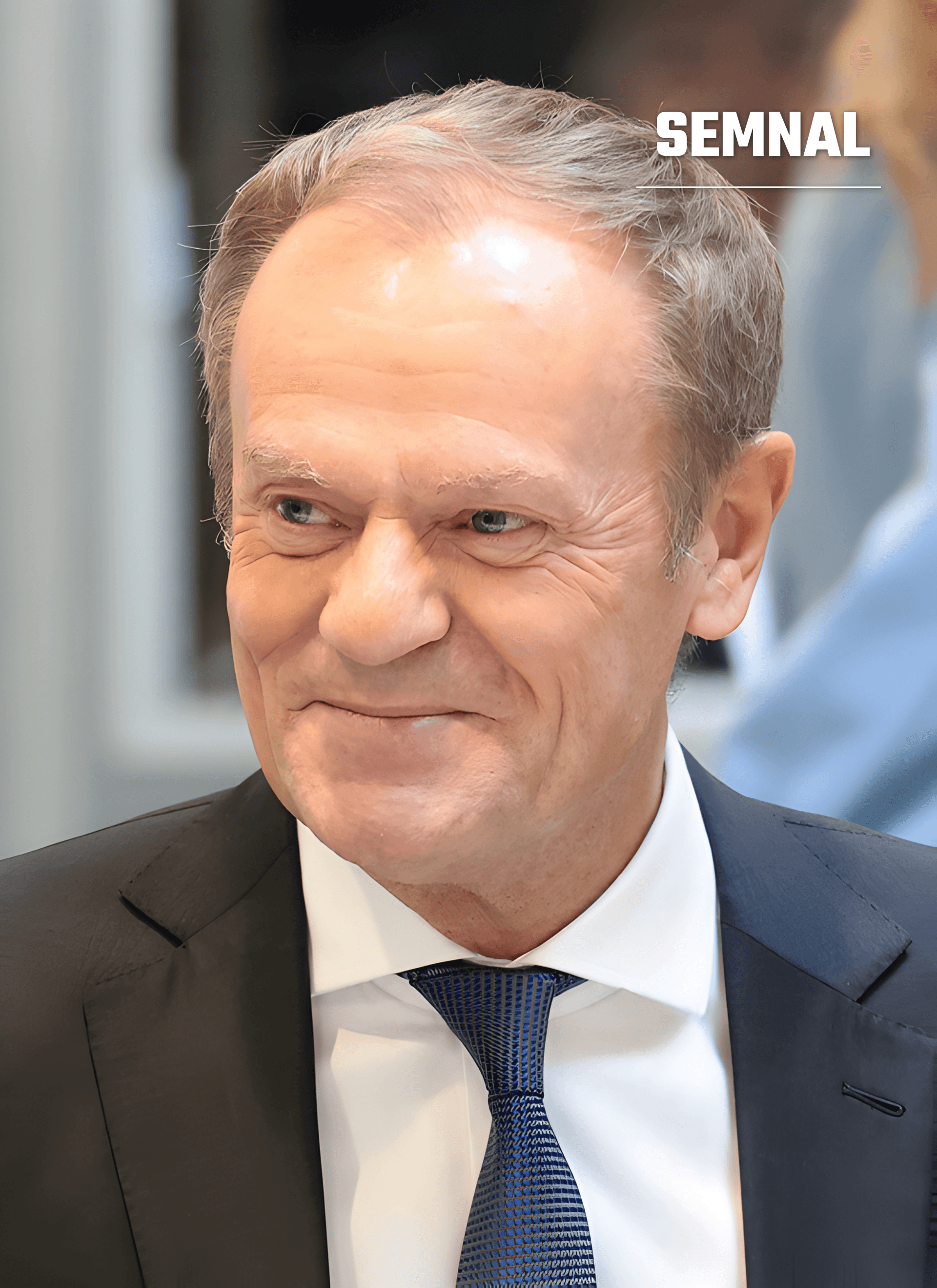 Donald Tusk, zâmbind ușor și privind lateral, cu părul gri, costum închis la culoare, cămașă albă și cravată albastră, într-un fundal estompat. (Foto: https://www.flickr.com/people/45198836@N04)