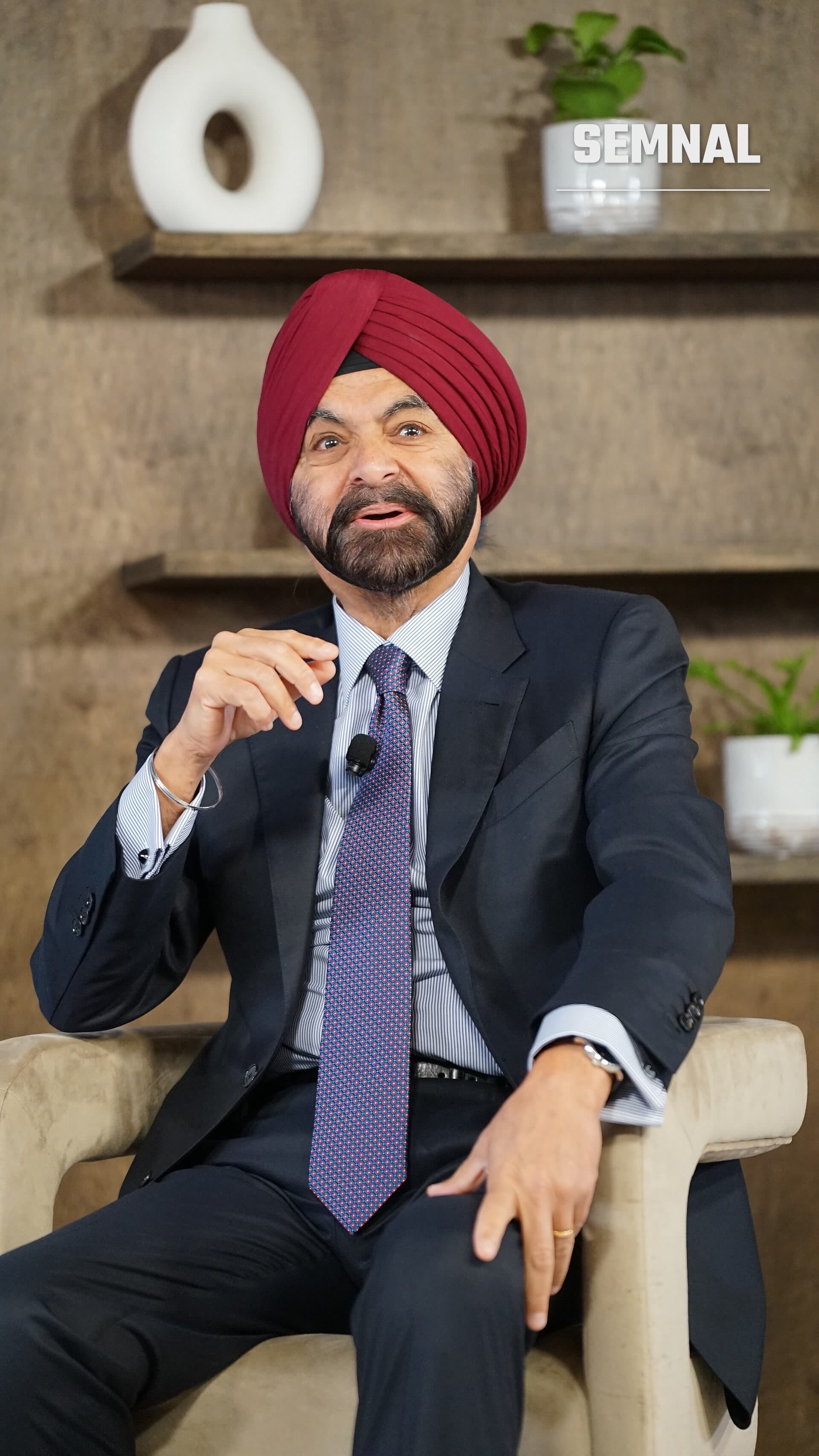 Ajay Banga, președintele Băncii Mondiale, ședea pe o canapă în timp ce vorbește, purtând turban roșu și costum negru cu cravată albastră cu puncte, într-un cadru interior modern. (Foto: Xuthoria)