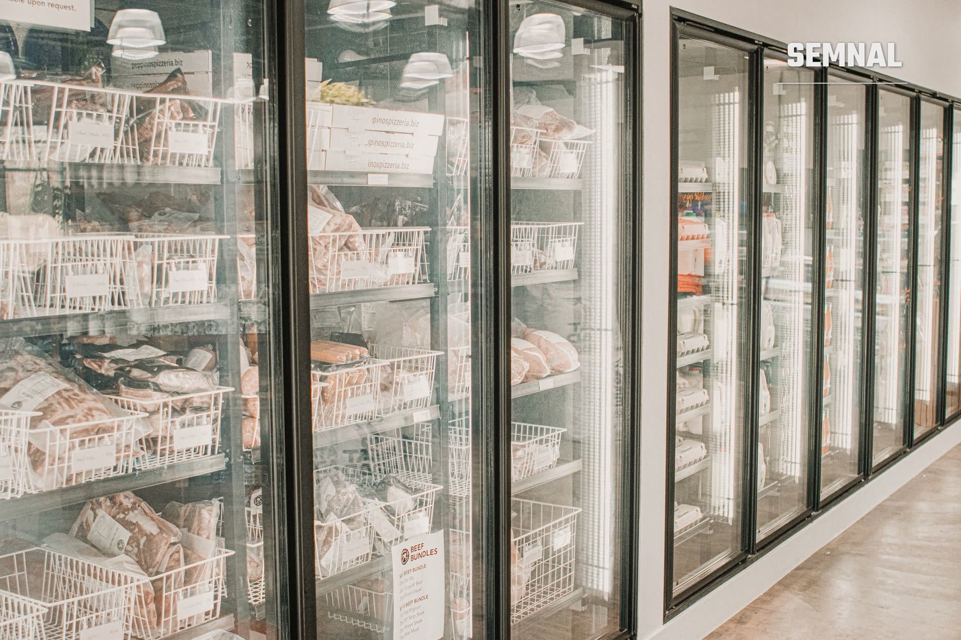 Vitrine frigorifice cu ușile din sticlă și cadre metalice conținând produse alimentare în coșuri albe de plastic, dispuse într-un rând în mediul unui supermarket modern. (Foto: Daniel & Hannah Snipes / Pexels)