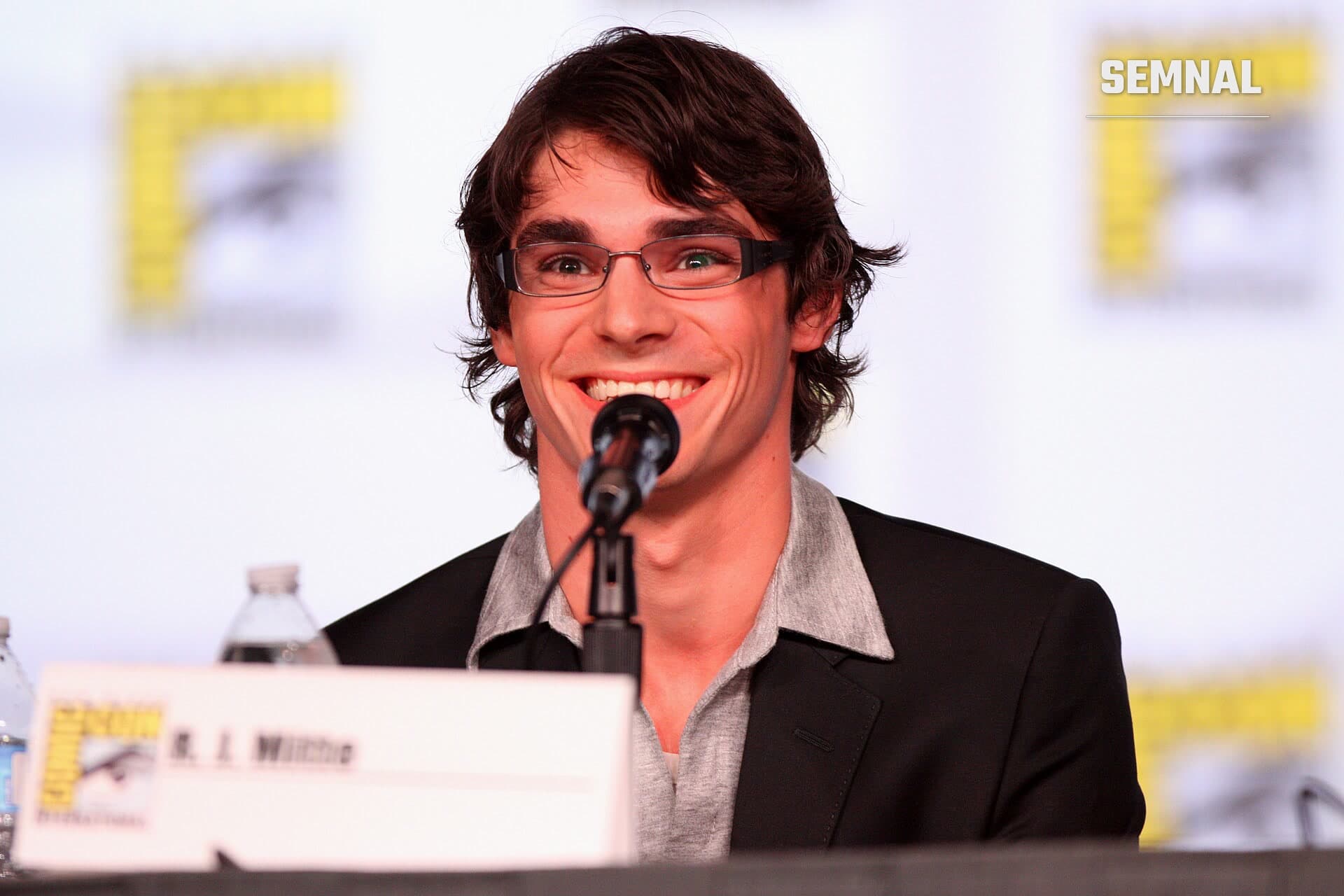 RJ Mitte zâmbind în fața unui microfon, la o masă de conferință; poartă ochelari cu ramă subțire și sacou negru peste o cămașă gri (Foto: Gage Skidmore from Peoria, AZ, United States of America)