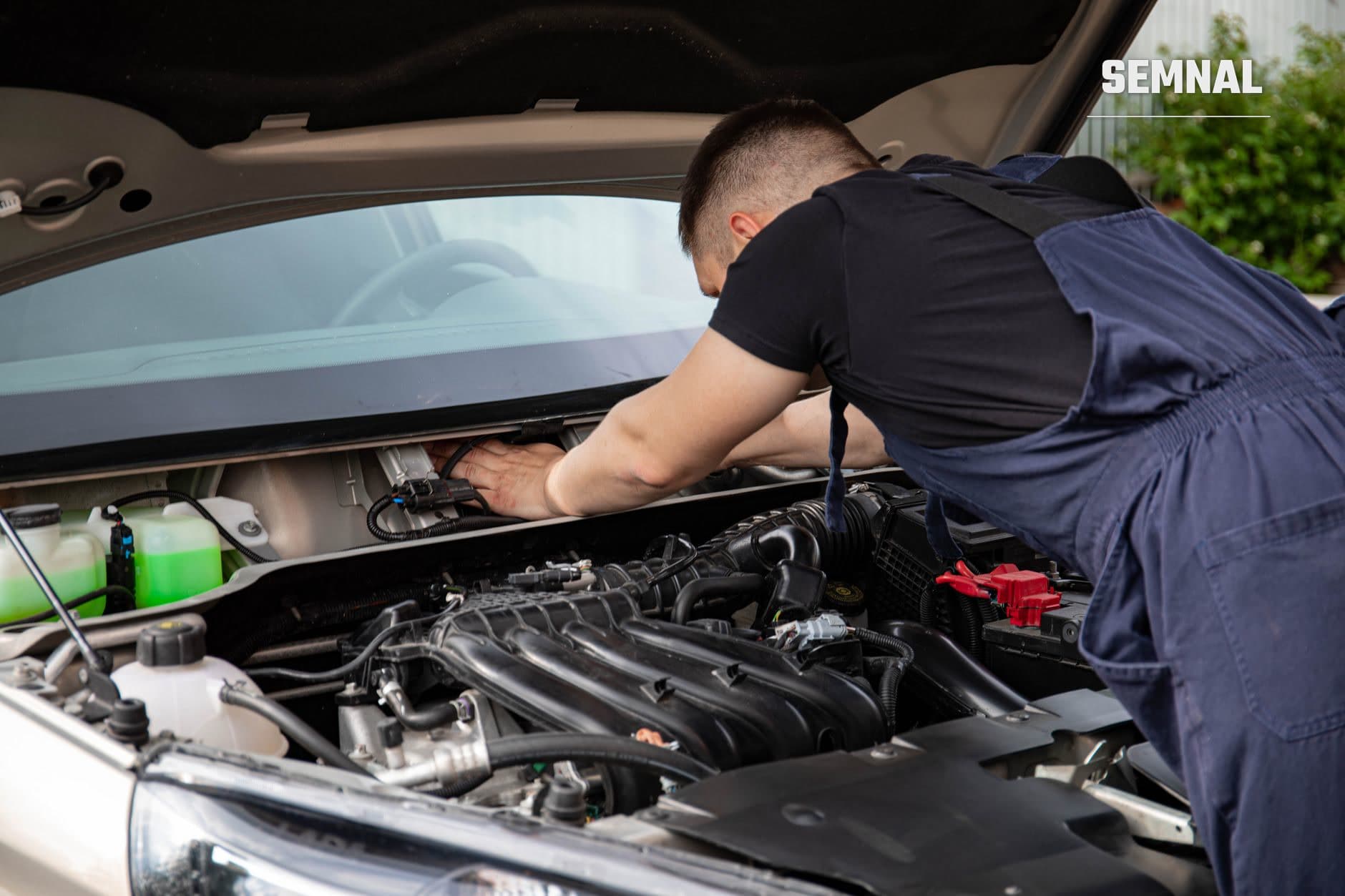 Tehnician în uniformă de lucru inspectând și întreținând motorul unui automobil, cu capota ridicată și componente mecanice vizibile. (Foto: Sergey Meshkov / Pexels)