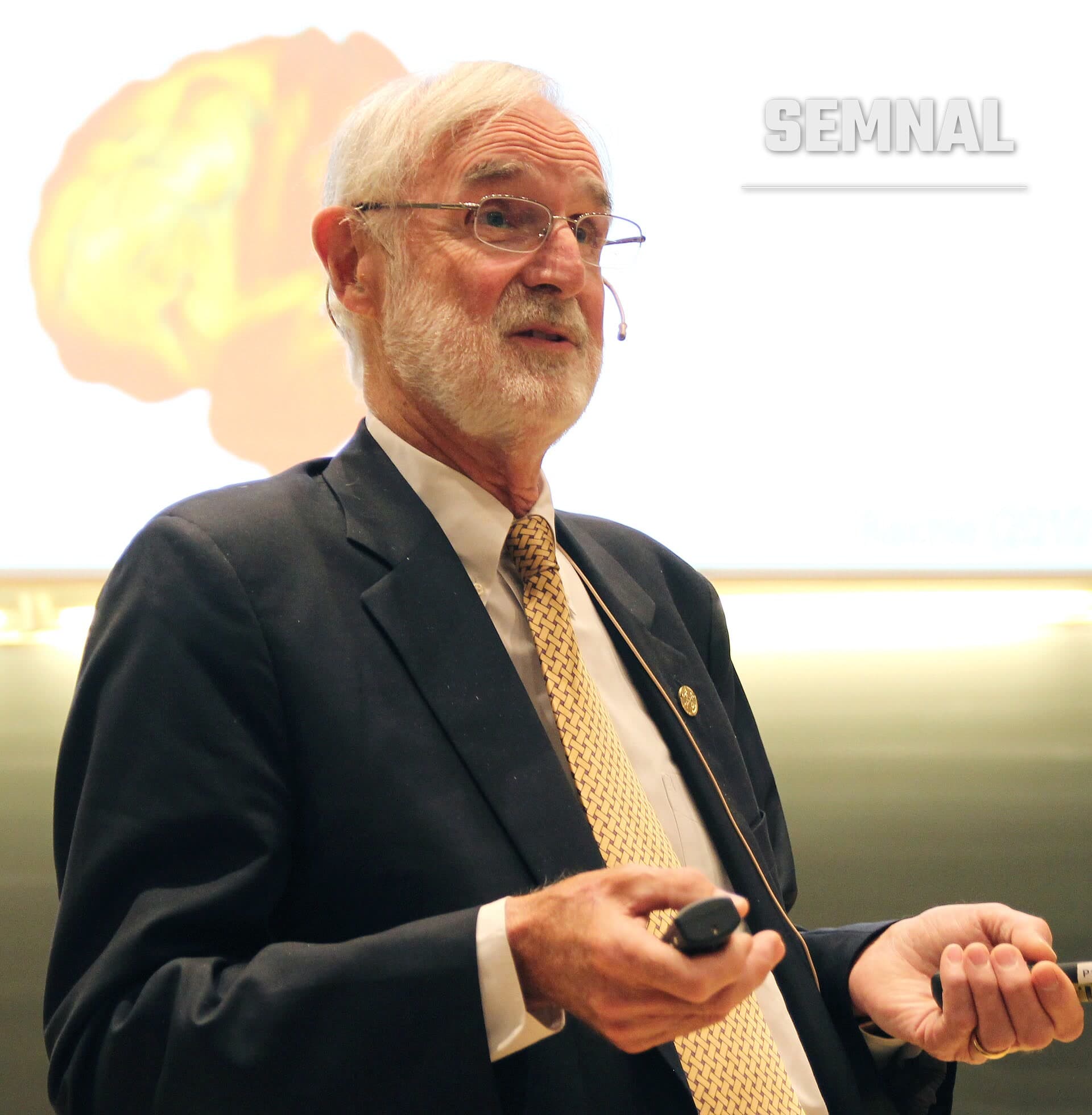 Marcus Raichle, neurolog, vorbind la o conferință, cu microfon pe cap și telecomandă în mână, în fața unui ecran cu imaginea unui creier. (Foto: Per Henning/NTNU)
