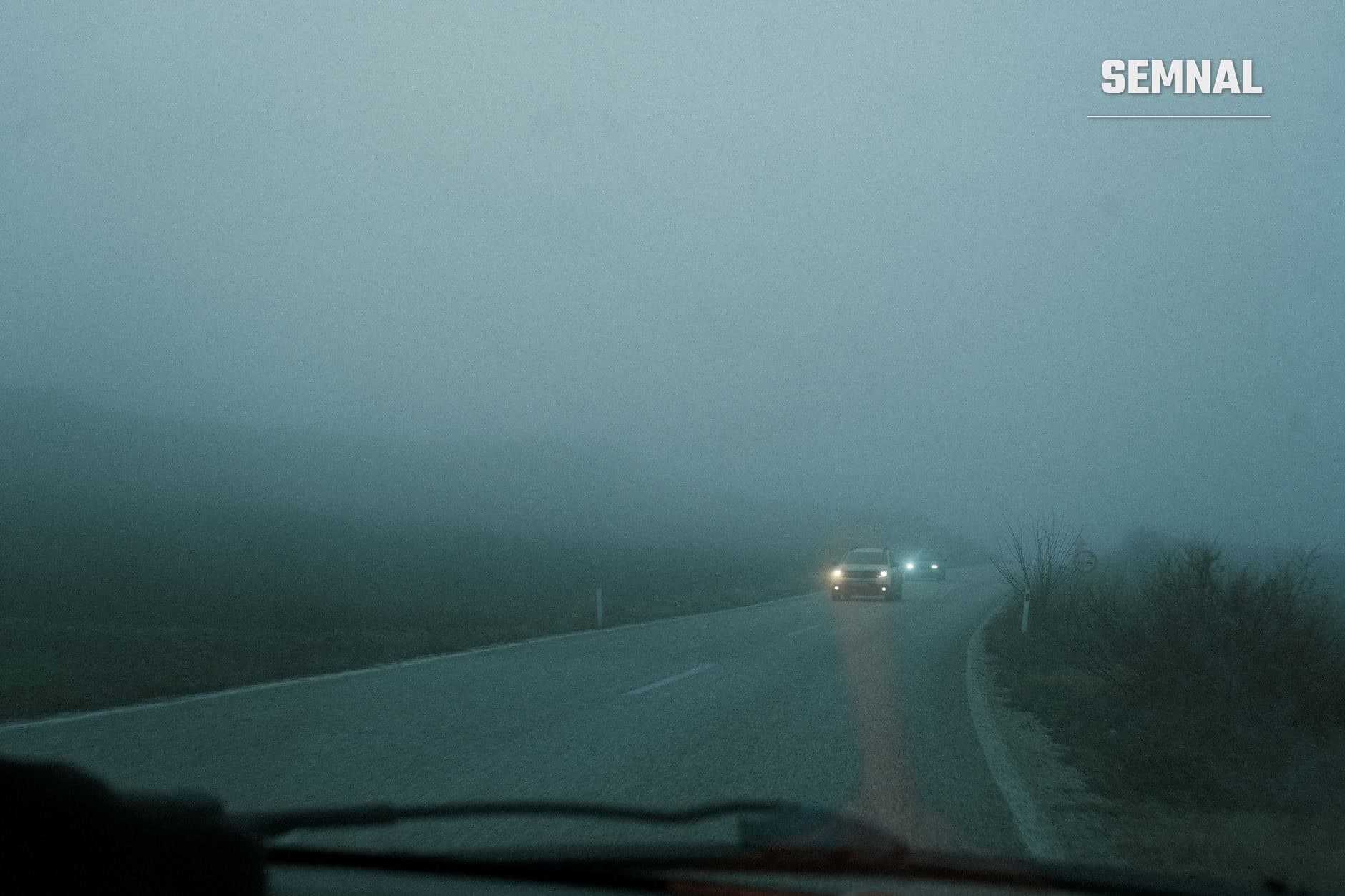Drum asfaltat învăluit în ceață densă, cu două autoturisme cu farurile aprinse, văzut din interiorul unei mașini (Foto: Alican Helik / Pexels)