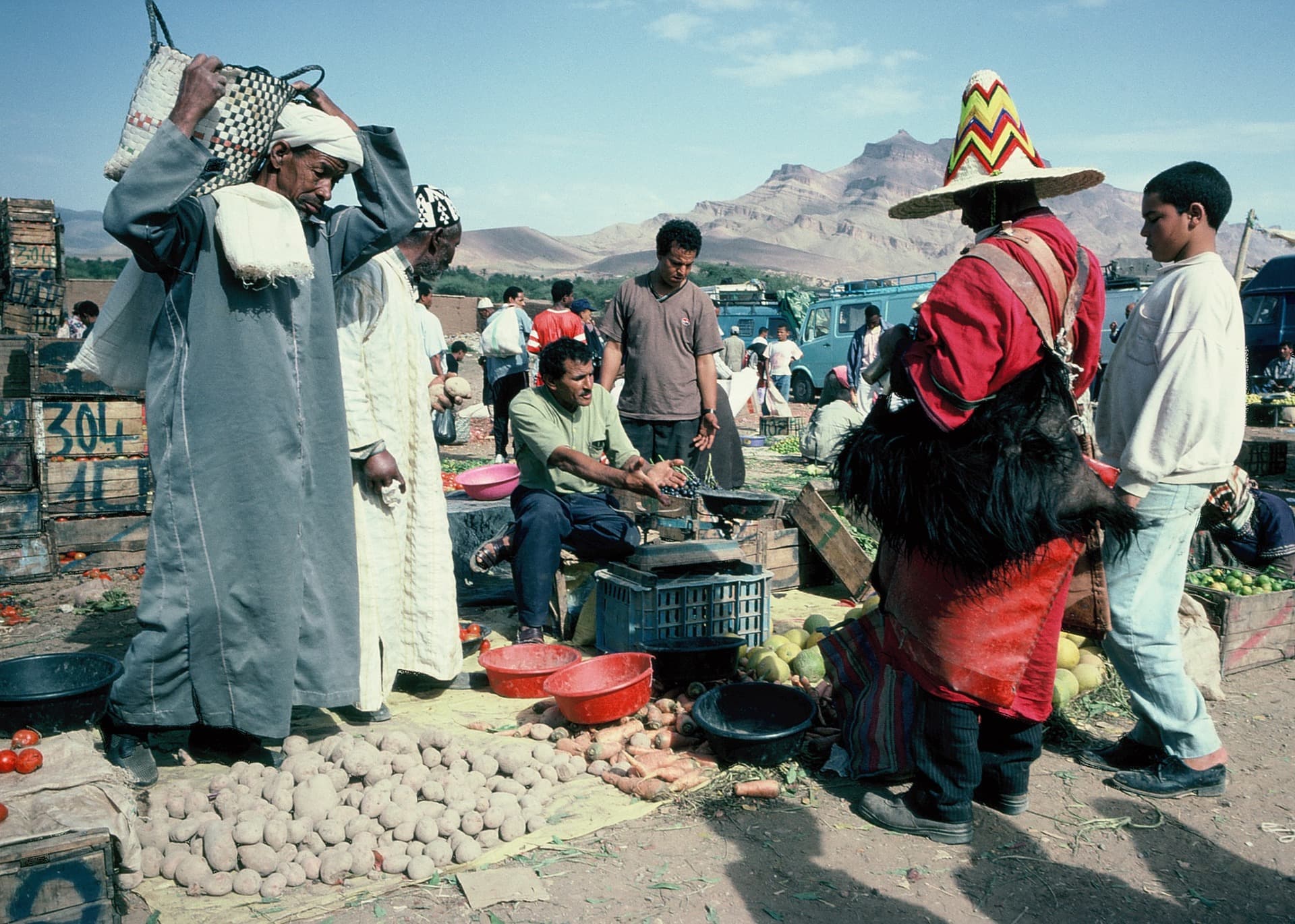 Piață în aer liber într-o localitate rurală din Maroc: vânzători de cartofi, morcovi și roșii, cumpărători și munți în fundal