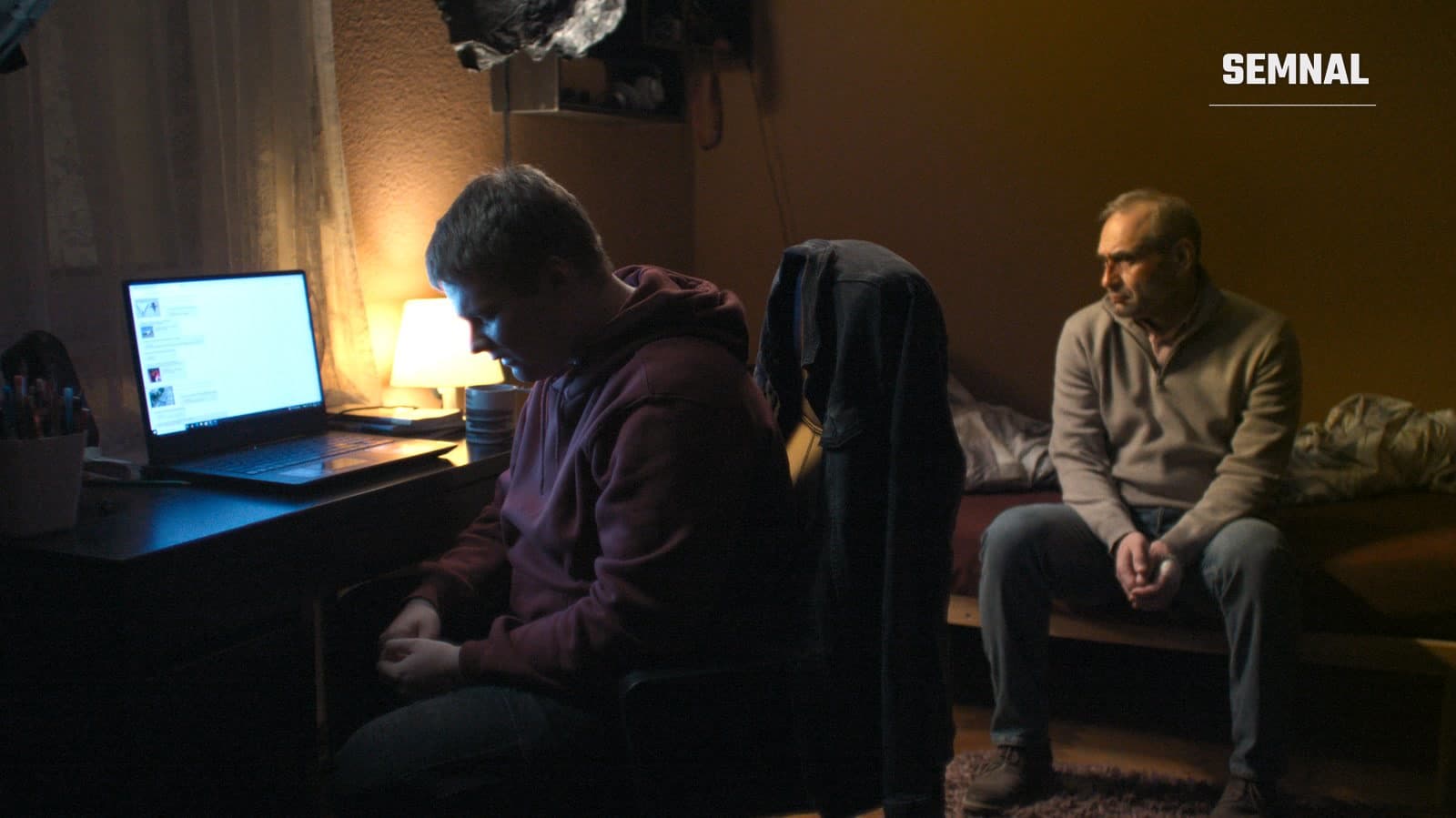 Scenă din filmul „Nu e locul tău aici”: un adolescent stă la birou, în fața unui laptop, în timp ce tatăl lui, așezat pe pat, îl privește îngrijorat.