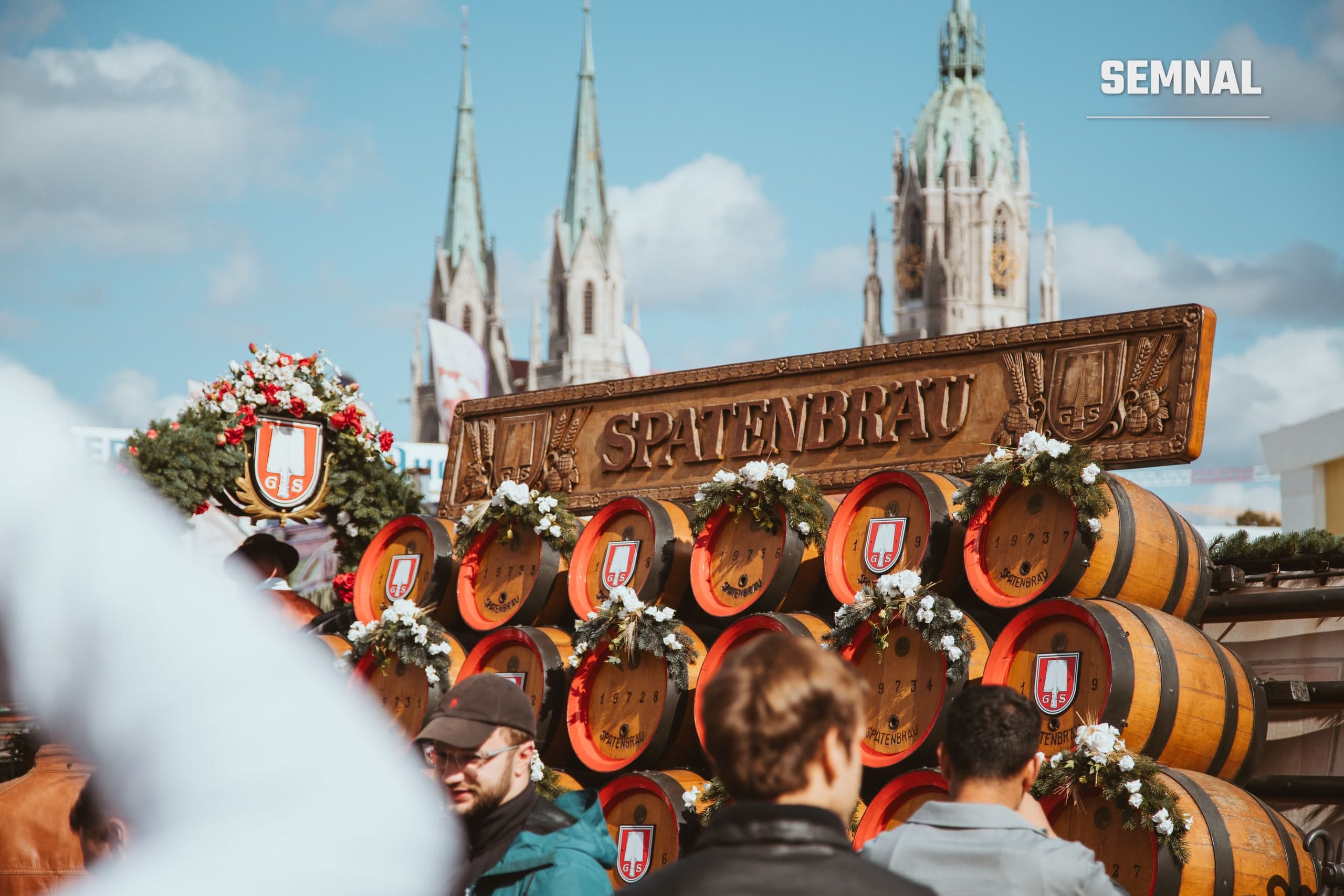 Butoaie de bere decorate în fața cortului Spatenbräu de la Oktoberfest din München, cu vizitatori în costume bavareze tradiționale.