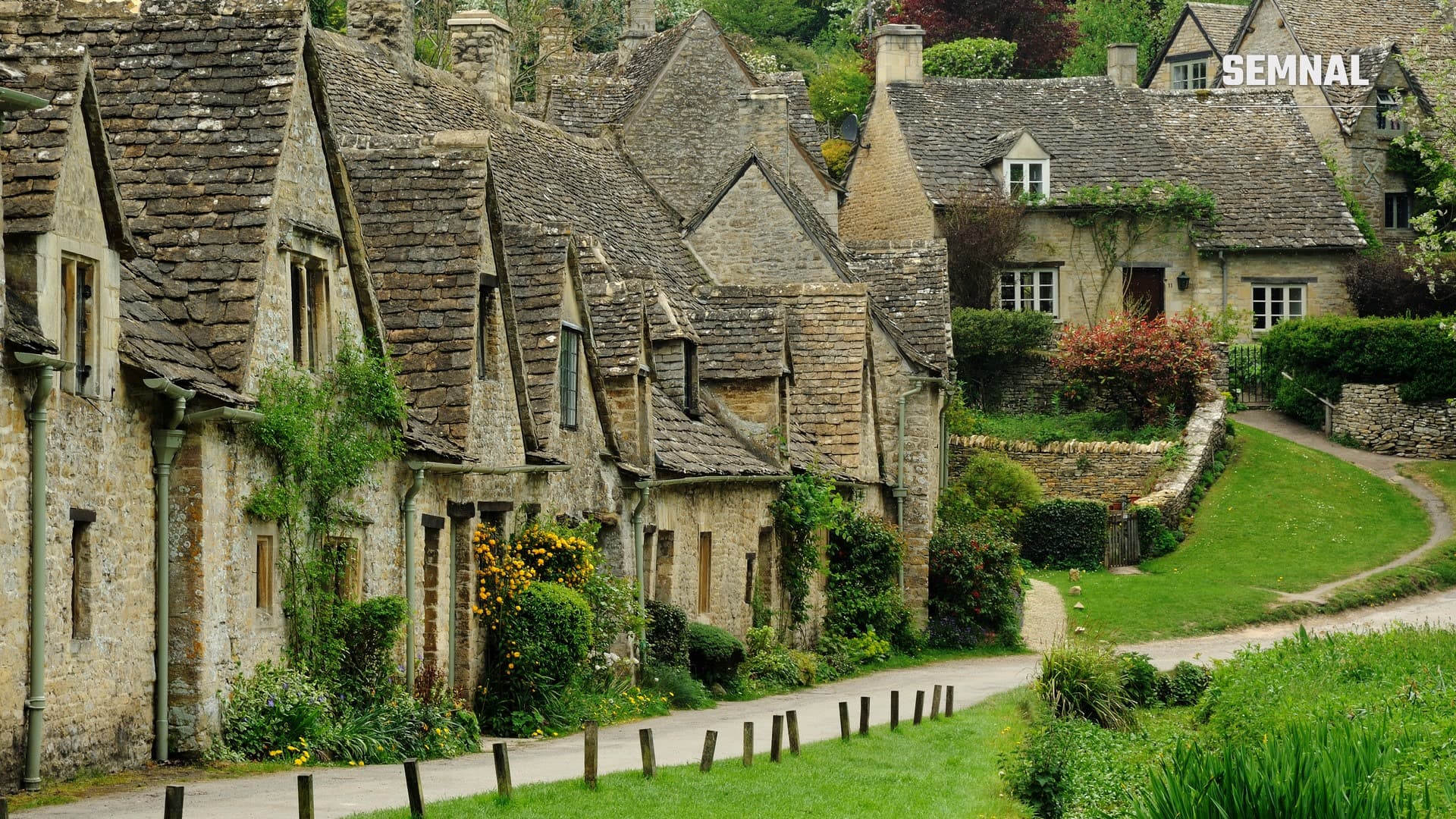 Arlington Row din satul Bibury, Anglia: căsuțe din piatră aurie cu acoperișuri înclinate, aliniate de-a lungul unei alei pietonale înverzite