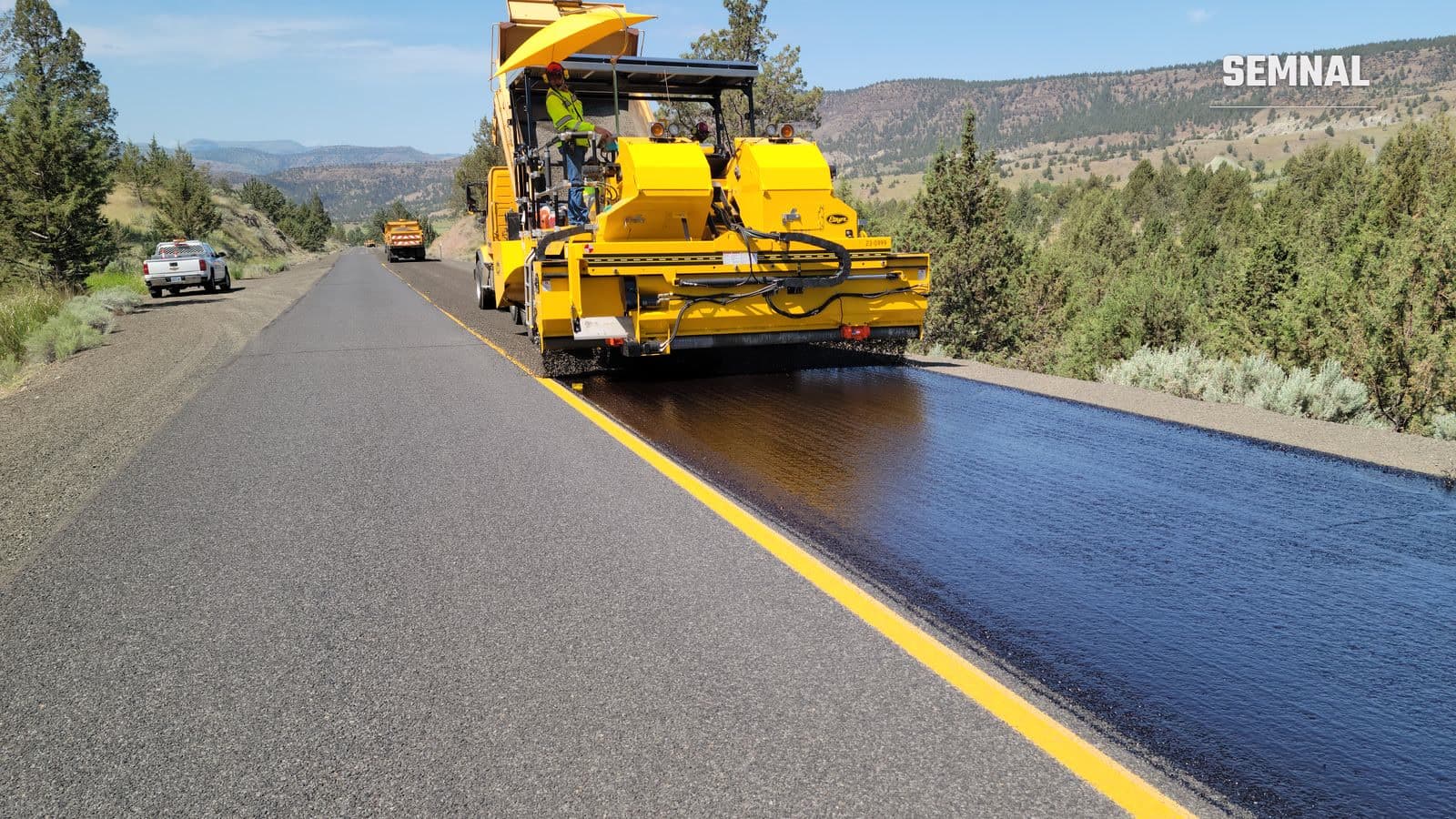 Muncitor în echipament de protecție supraveghează un utilaj galben care așterne asfalt pe un drum ce traversează un peisaj deluros, ilustrativă pentru lucrări de infrastructură rutieră