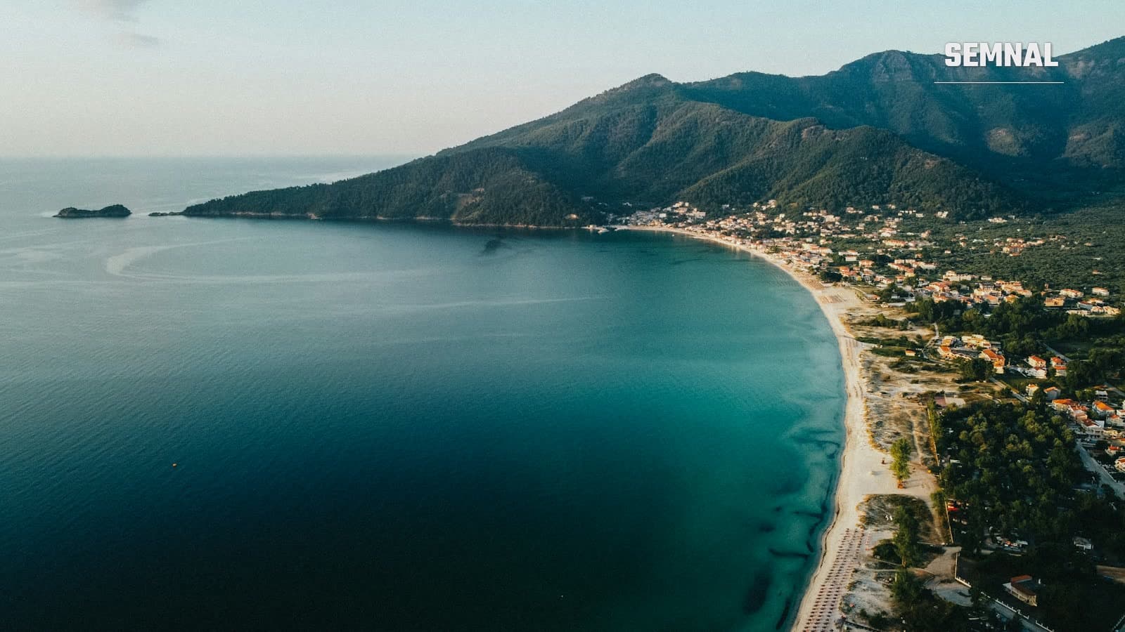 Vedere aeriană a unui golf din Thassos, cu plajă de nisip, sat construit pe malul mării și munți împăduriți în fundal