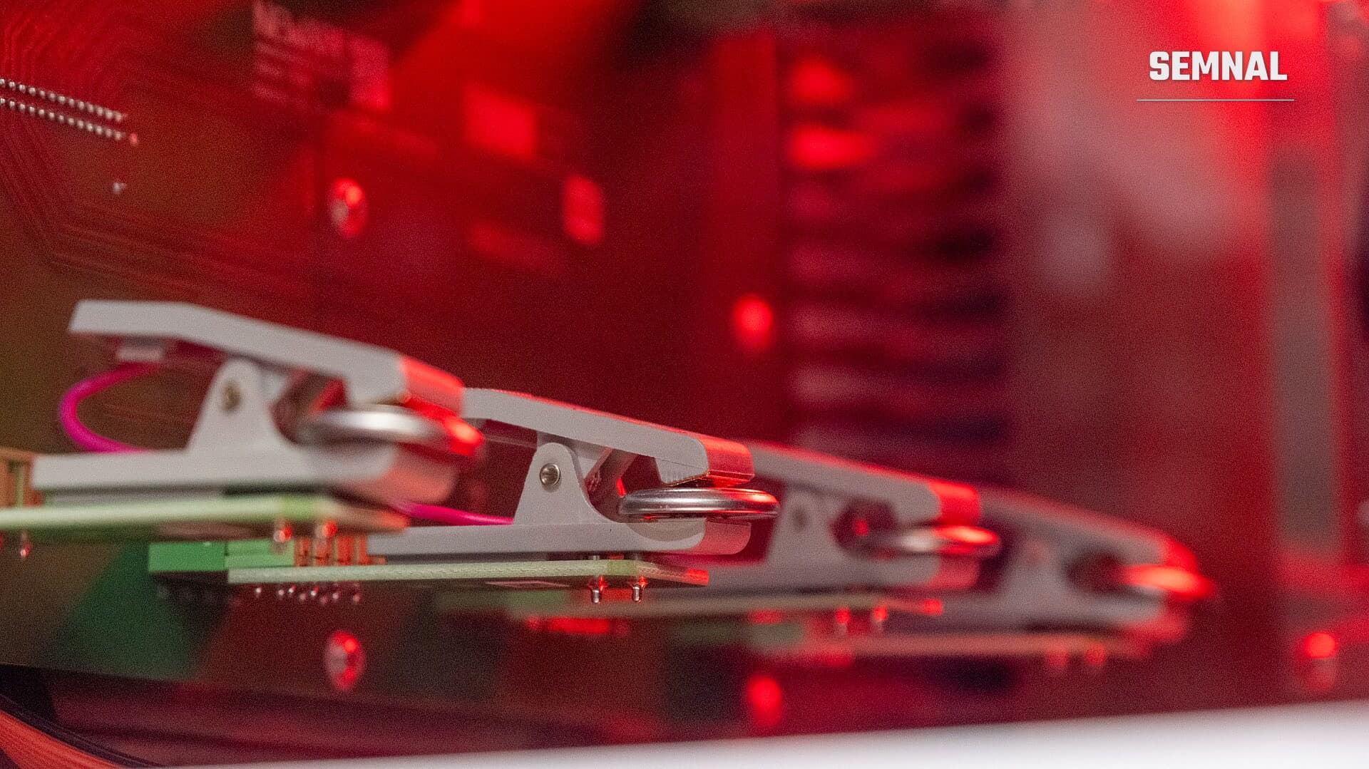 Cleme gri de testare prinse pe plăci electronice verzi, într-un dispozitiv de laborator pentru baterii, în lumină roșie