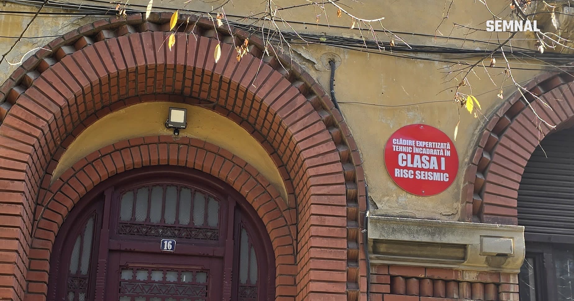 Intrarea clădirii de pe Strada Londra 16 din București: ușă de lemn sub un arc de cărămidă și, alături, panoul roșu „Clădire expertizată tehnic încadrată în clasa I risc seismic”