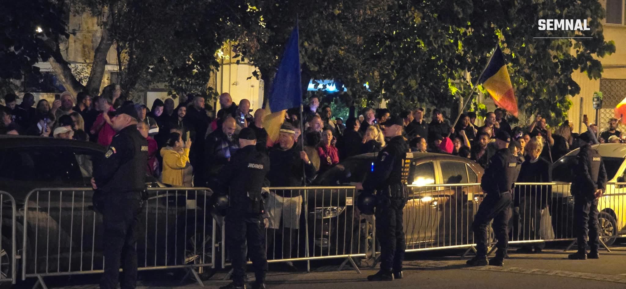 Oameni adunați noaptea în spatele unor garduri metalice, cu drapele ale României, păziți de jandarmi în uniformă închisă la culoare.