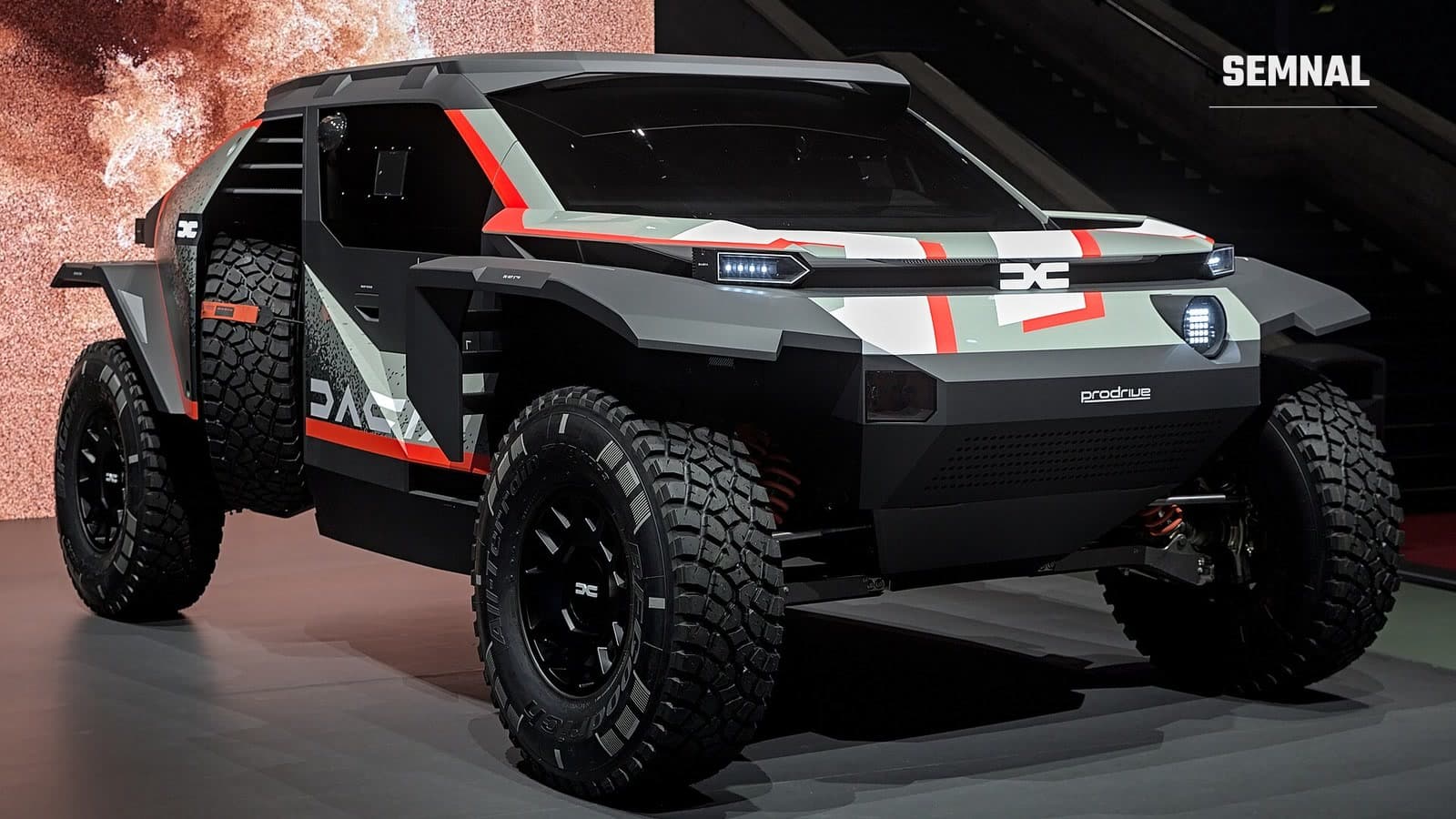 Prototipul de raliu-raid Dacia Sandrider, cu caroserie gri și neagră cu accente roșii, anvelope off-road late și sigla Dacia pe botul mașinii, expus într-un hol de expoziție
