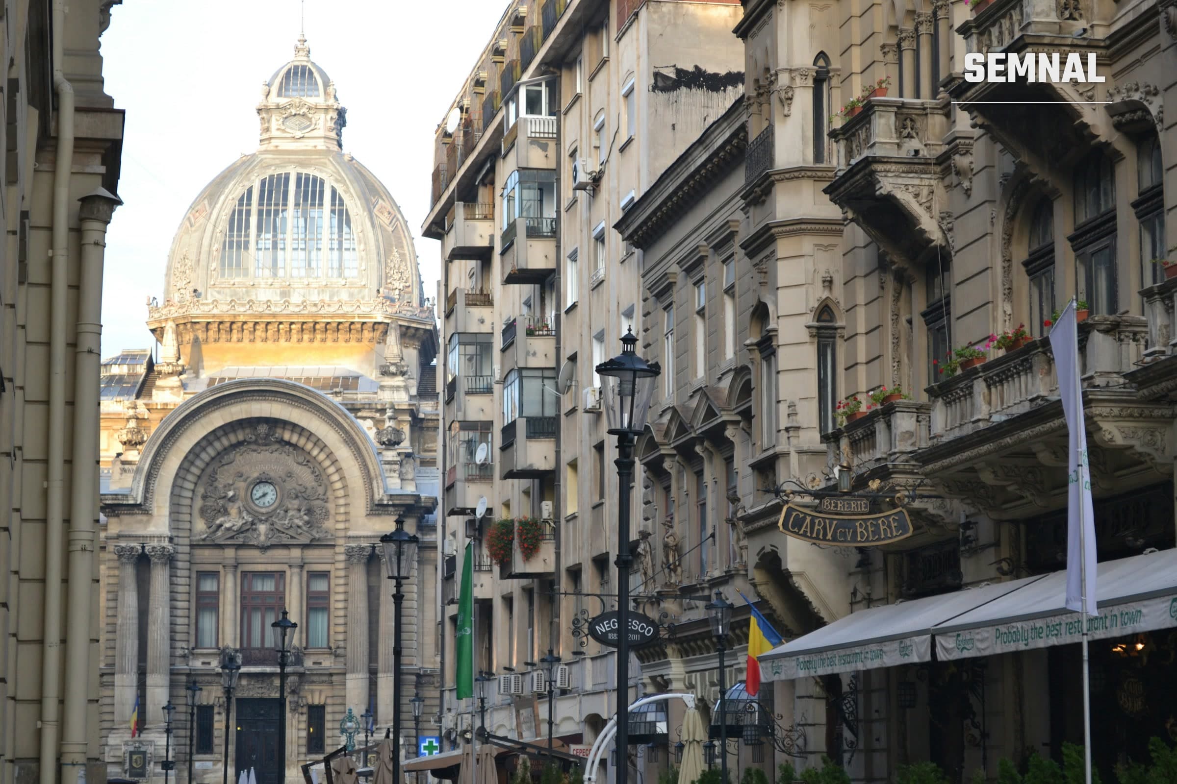 Stradă din centrul Bucureștiului cu clădiri de la începutul secolului XX, felinare și firma berăriei Caru' cu Bere, cu cupola Palatului CEC în fundal