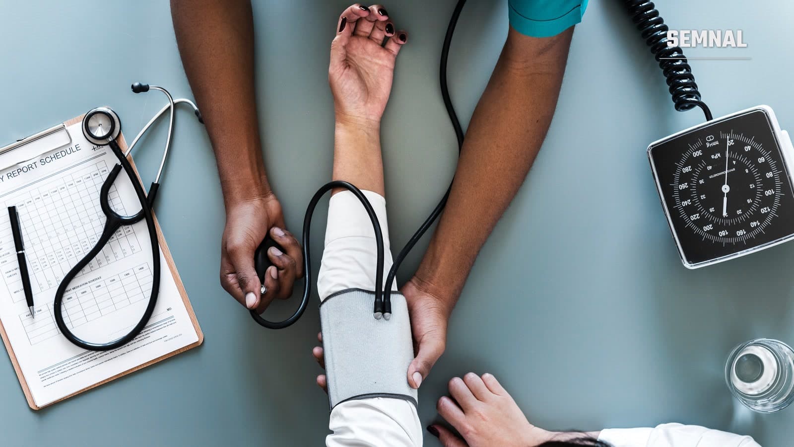 Mâinile unui medic măsoară tensiunea arterială unui pacient, cu manșetă și tensiometru, lângă un stetoscop și un clipboard