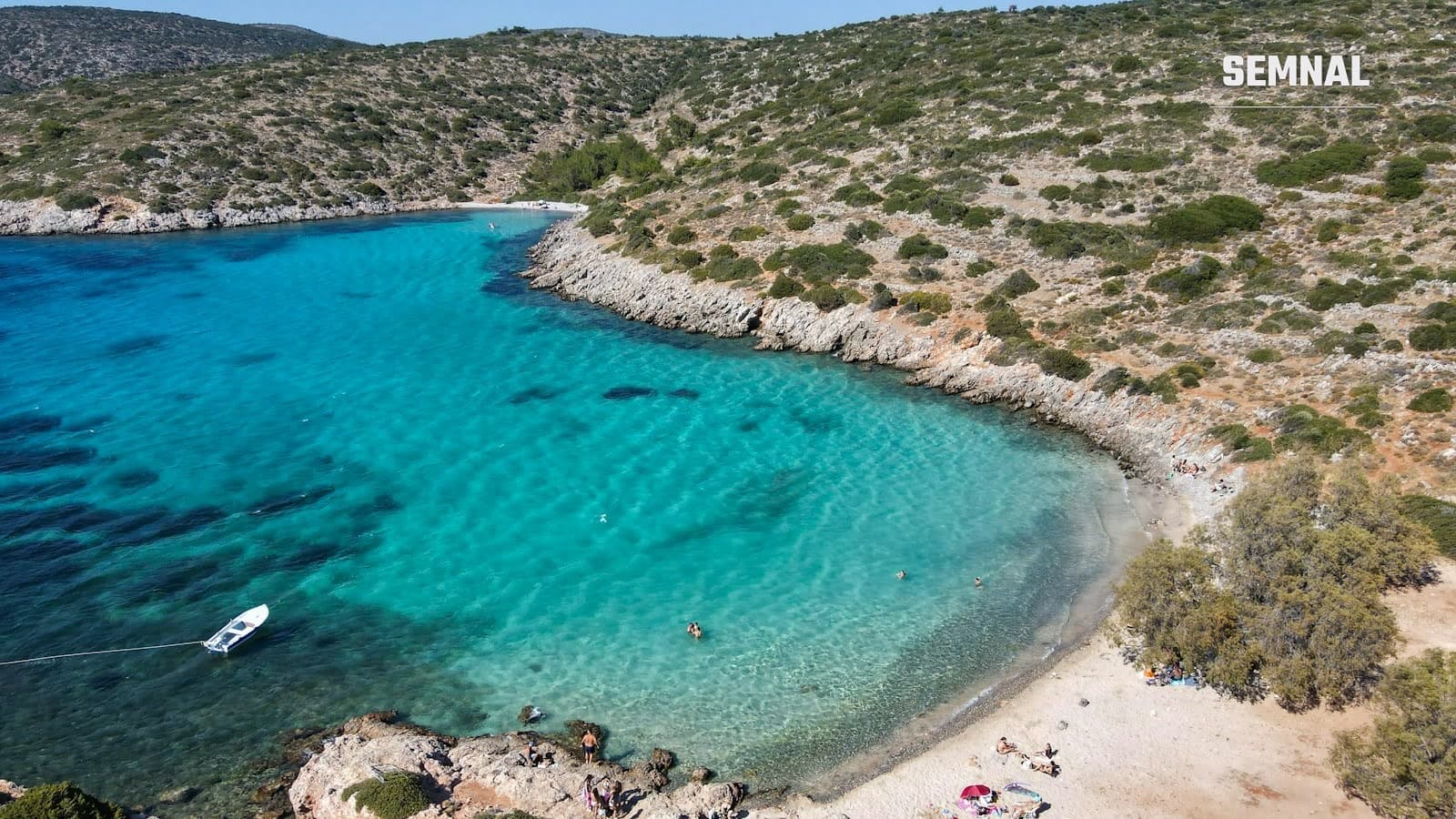 Golfuleț cu apă turcoaz pe coasta insulei Chios, la plaja Agia Dynami, văzut din dronă, cu o barcă albă ancorată și câțiva înotători