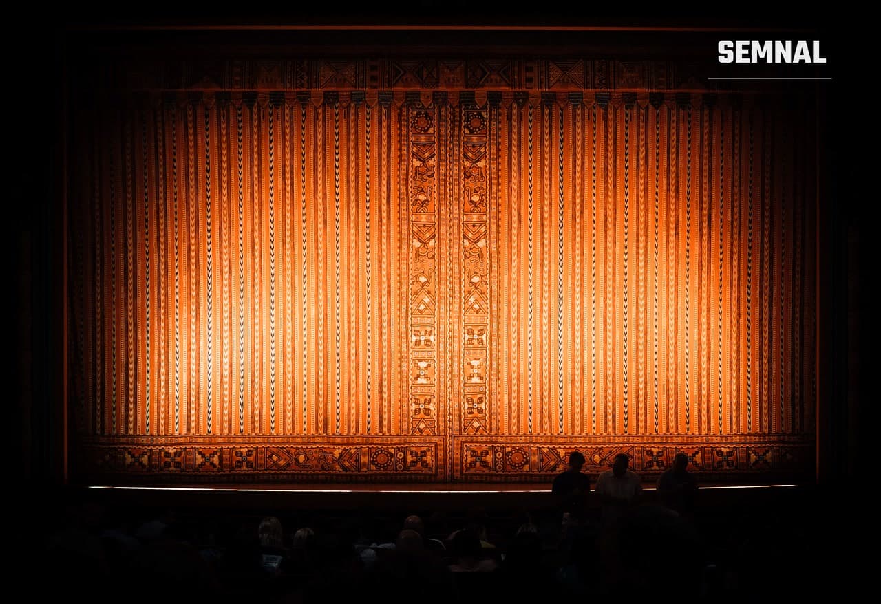 Cortina luminată a unei scene de teatru, cu siluetele spectatorilor din sală