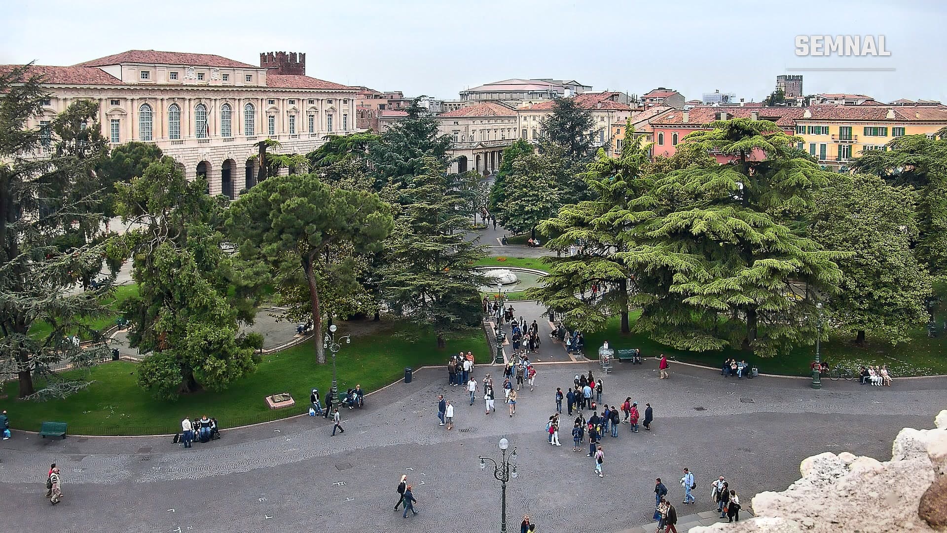 Turiști pe pavajul din Piazza Bra, Verona, între grădina cu pini și cedri și clădirile istorice din jur, văzuți de pe Arena