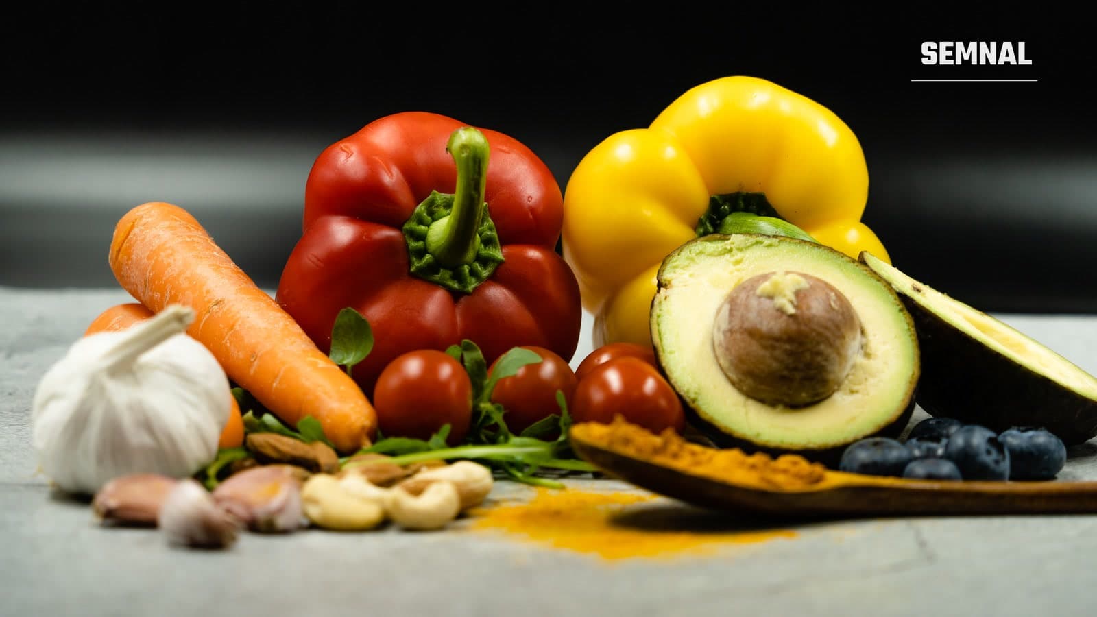 Ardei roșu și galben, morcov, roșii cherry, usturoi, avocado tăiat, afine, caju și migdale, aranjate pe o suprafață gri, pe fundal întunecat
