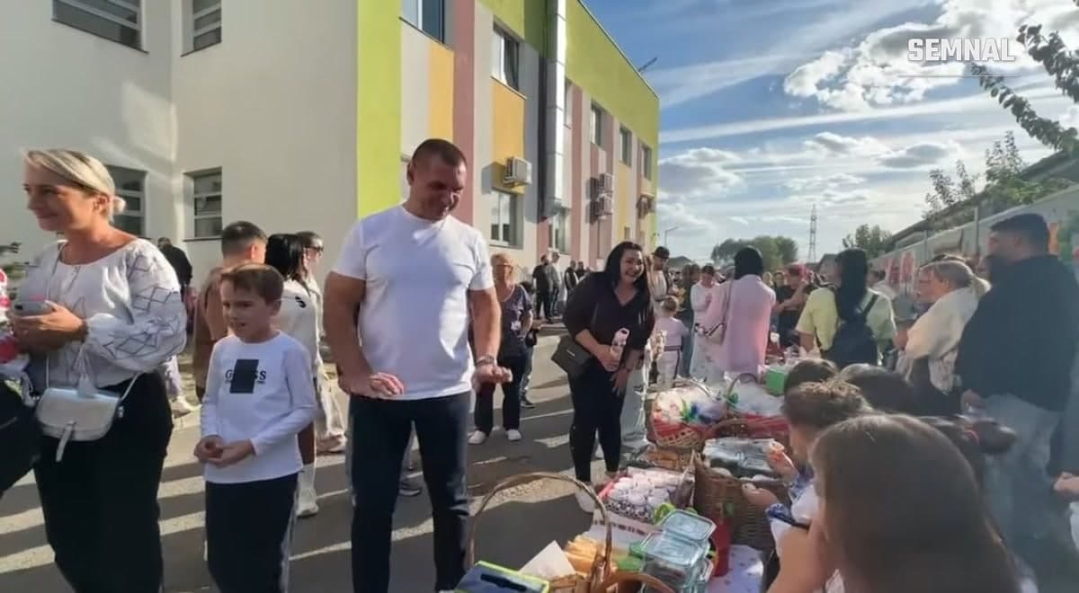 Părinți și copii în jurul unei mese cu coșuri de produse de toamnă, în fața unei clădiri cu fațadă colorată, la Festivalul Toamnei
