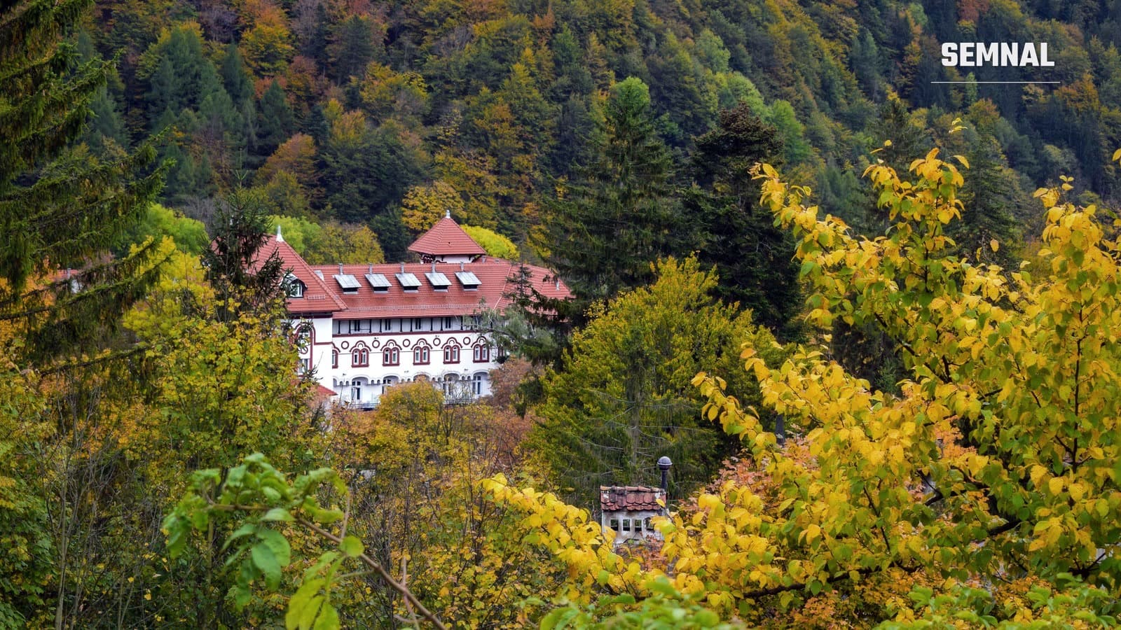 Clădire albă cu acoperiș roșu printre arbori cu frunze galbene și portocalii, într-o pădure de toamnă din Sinaia, pe Valea Prahovei