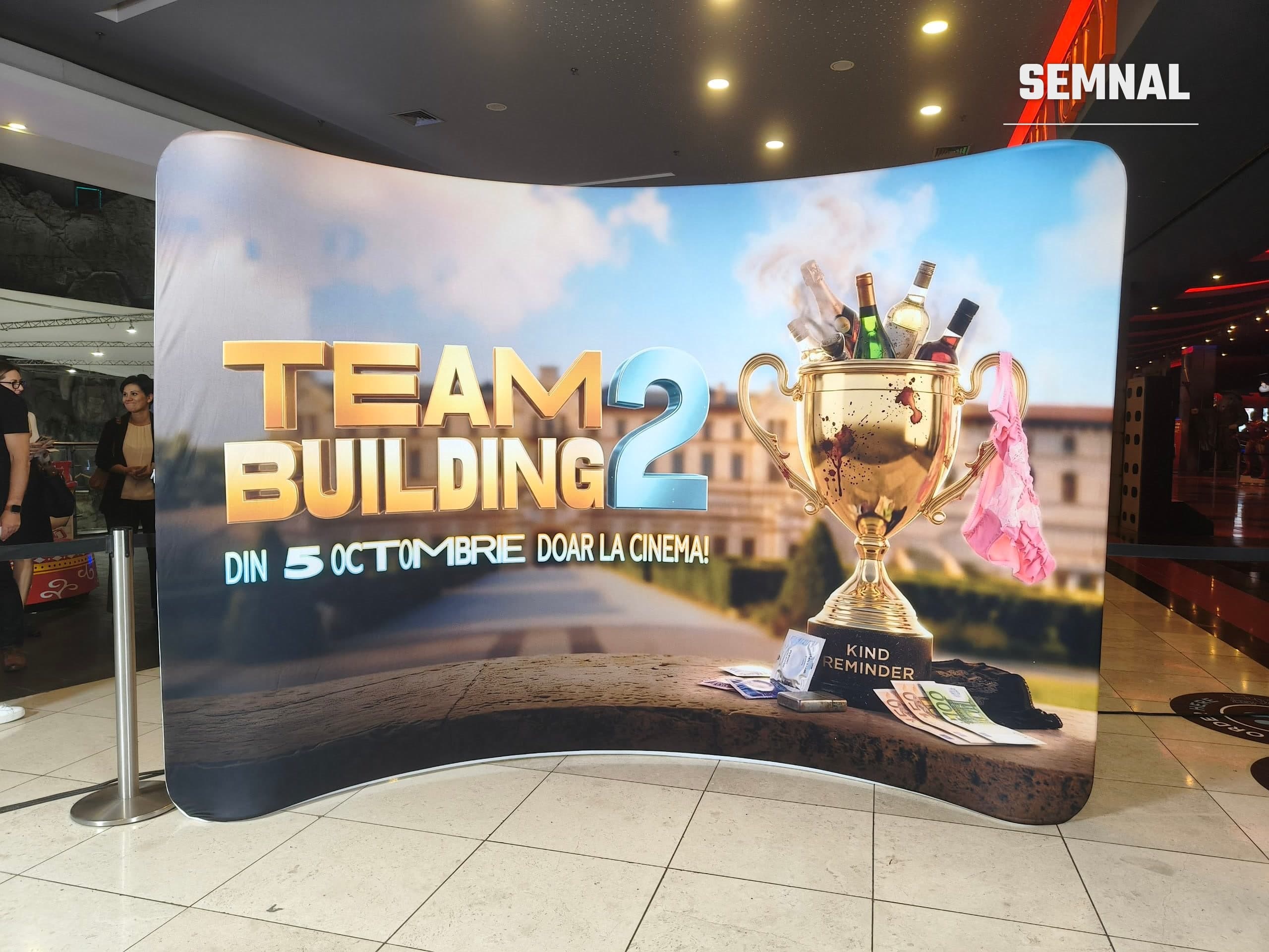 Panou promoțional Teambuilding 2 în holul Cinema City AFI Cotroceni, cu un trofeu auriu plin de sticle și anunțul lansării din 5 octombrie