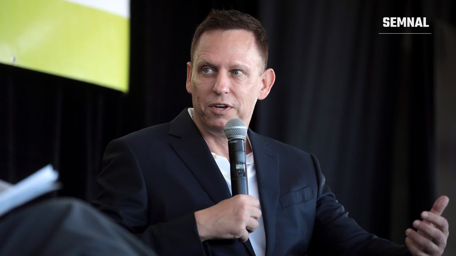 Peter Thiel vorbind la microfon pe o scenă, în costum închis la culoare, cu un panou verde și o cortină neagră în fundal
