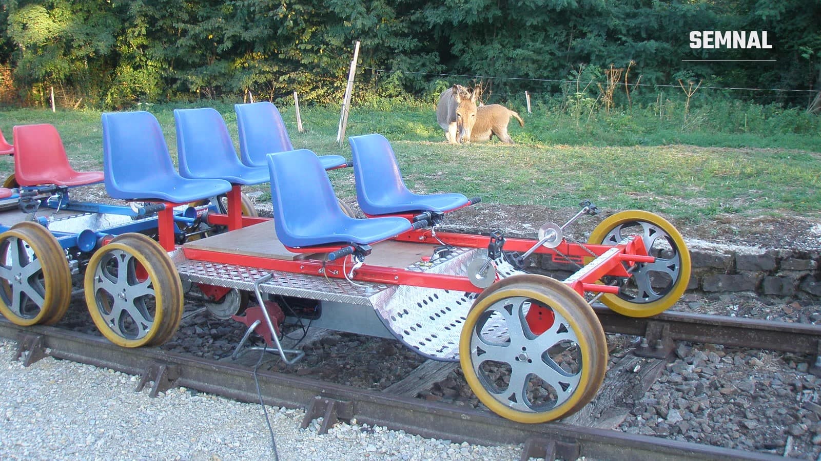 Velorail cu scaune albastre și roșii, pe o cale ferată îngustă, într-un cadru rural cu iarbă și copaci, imagine ilustrativă din Portugalia